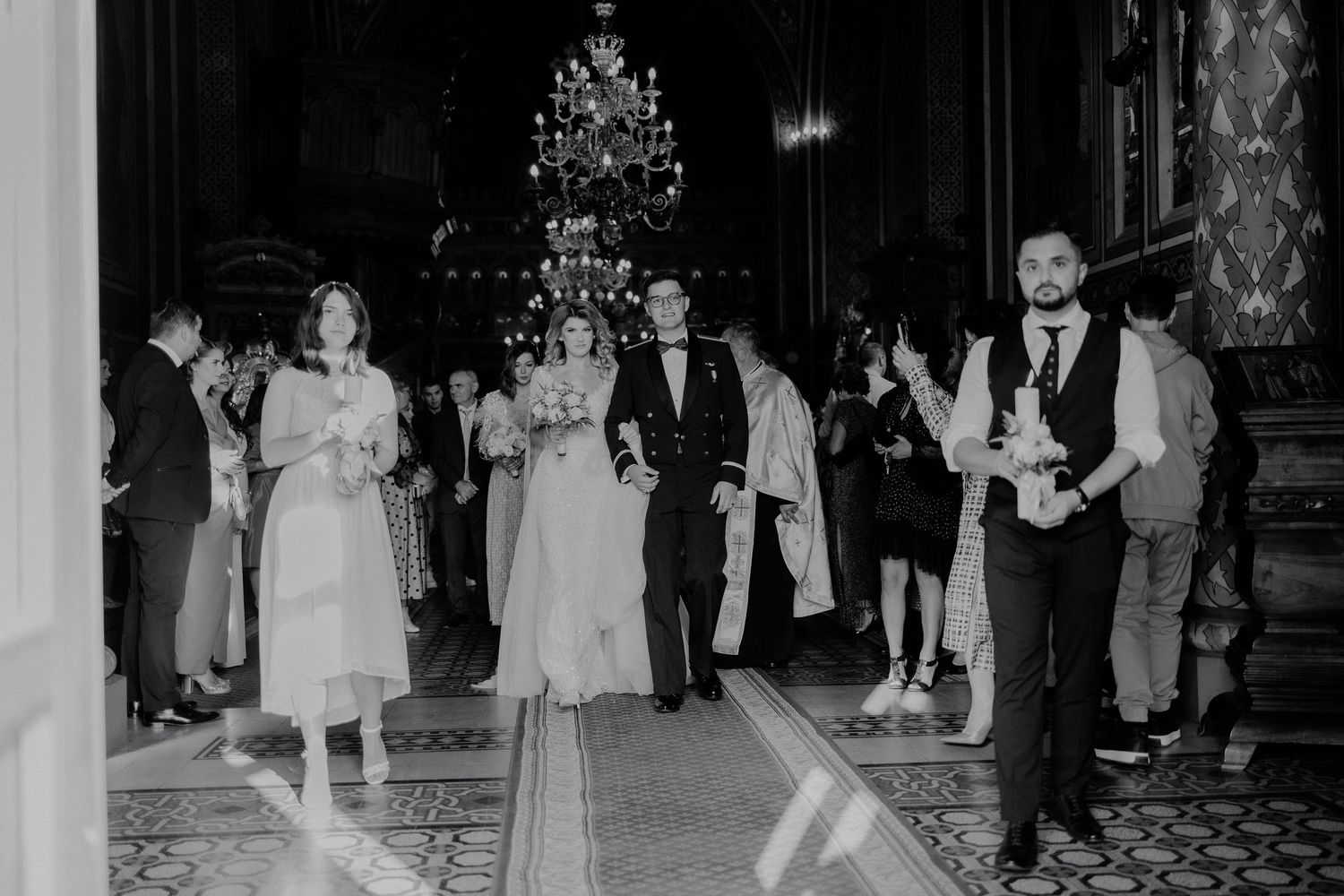 Miri pășind spre ieșirea din biserică la finalul ceremoniei; fotografie alb-negru surprinzând un moment solemn sub lumina candelabrelor.