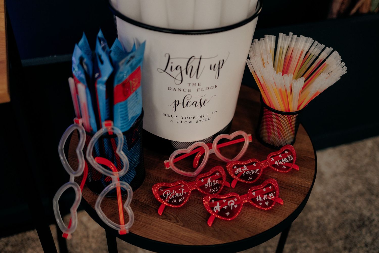 Ochelari în formă de inimă și bețișoare luminoase pe o măsuță; accesorii distractive pentru invitați cu mesajul „Light up the dance floor”.