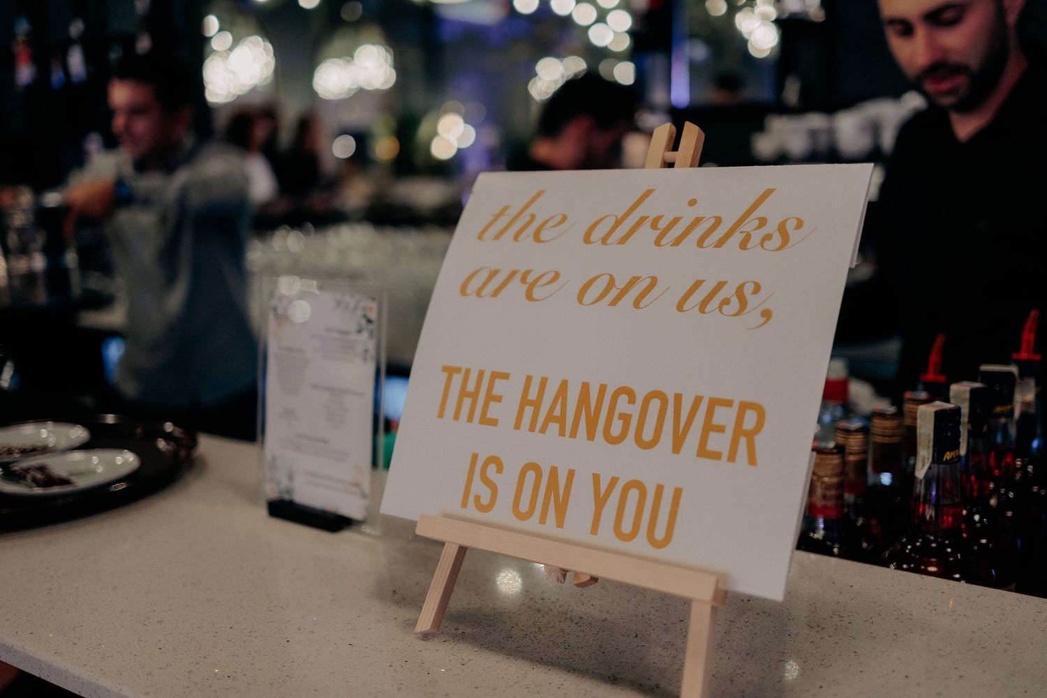 Semn haios la barul unei nunți cu mesajul „the drinks are on us, THE HANGOVER IS ON YOU”, așezat pe un șevalet mic de lemn.