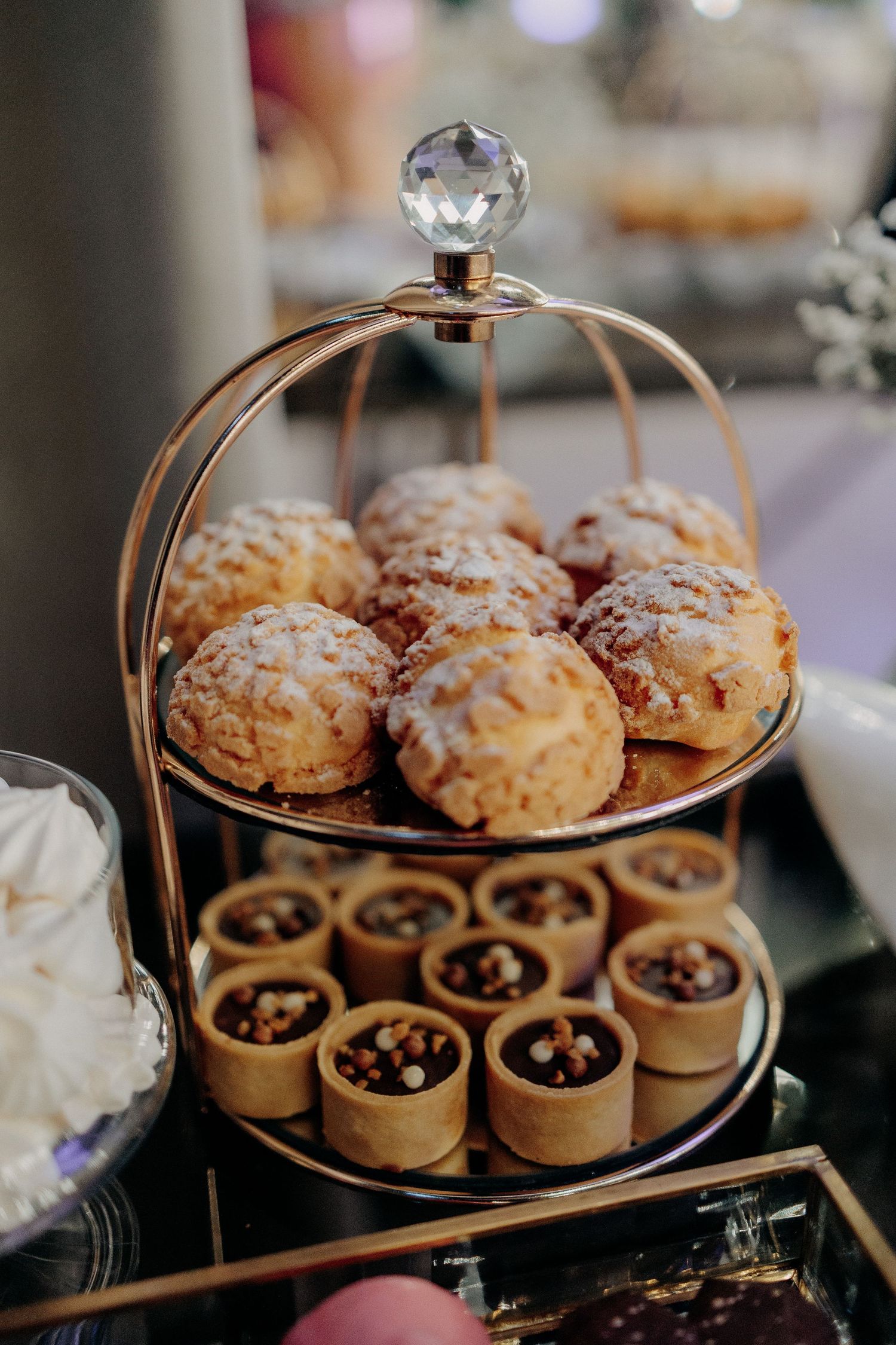 Mini tarte cu ciocolată și choux à la crème pudrate, aranjate elegant pe un suport auriu etajat la candy bar-ul unei nunți.
