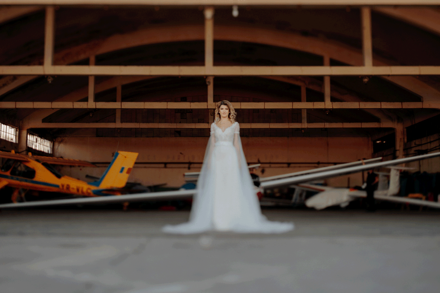 Mireasă stând elegant în centrul unui hangar de avioane; imagine dinamică ce alternează focalizarea între fundalul tehnic și silueta miresei