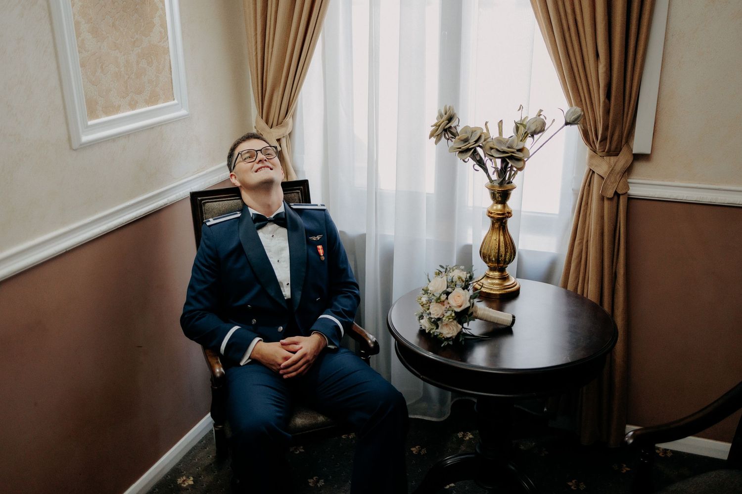 Portret relaxat cu mirele în uniformă militară, stând pe un scaun lângă o fereastră și privind în sus; buchetul miresei este așezat pe masă.