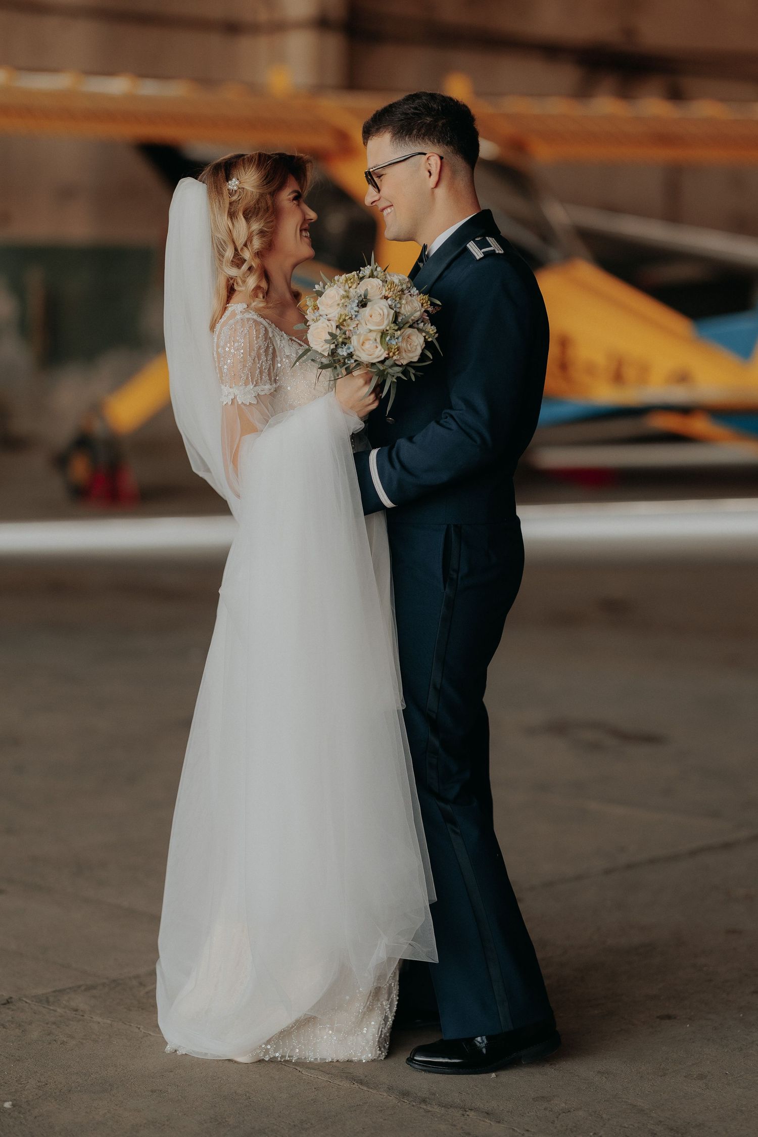 Miri zâmbindu-și cu dragoste într-un hangar, mireasa ținând un buchet elegant; cadru romantic lângă un avion galben, la sesiunea foto.