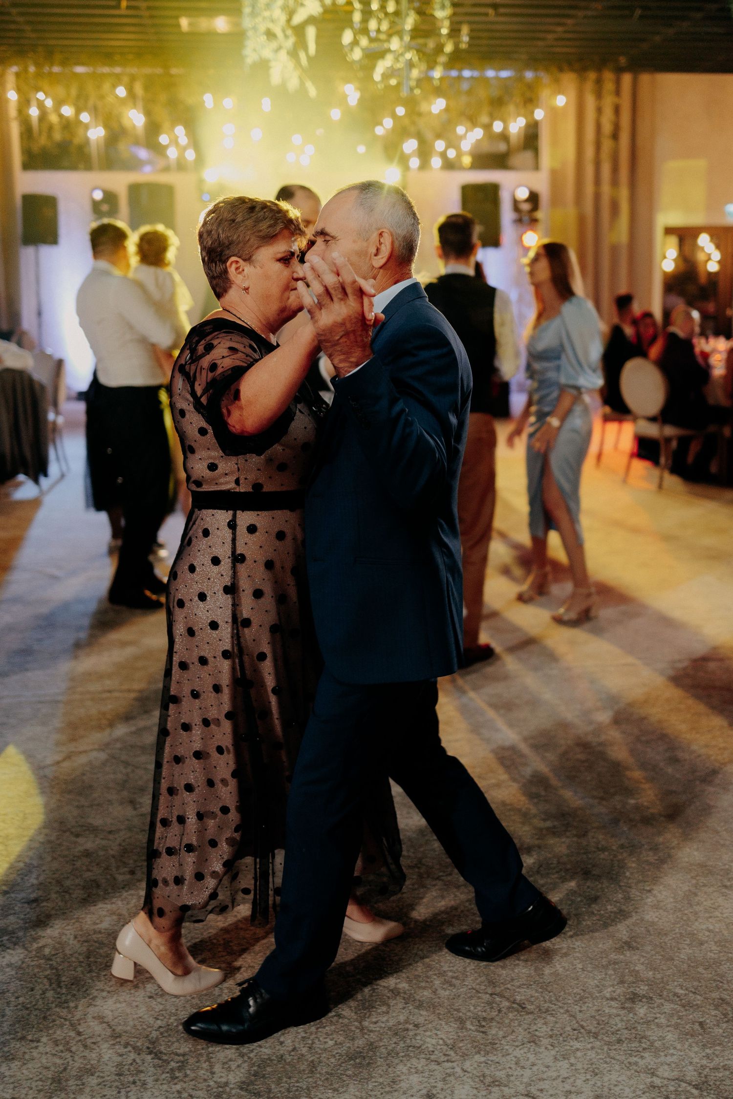 Cuplu în vârstă dansând elegant la o petrecere de nuntă; moment emoționant pe ringul de dans, sub lumini calde de sărbătoare.