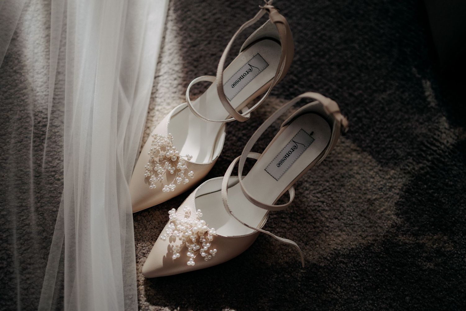 Plan detaliu cu pantofii de mireasă din satin bej, decorați cu perle delicate, așezați lângă vălul de mireasă în lumina naturală.