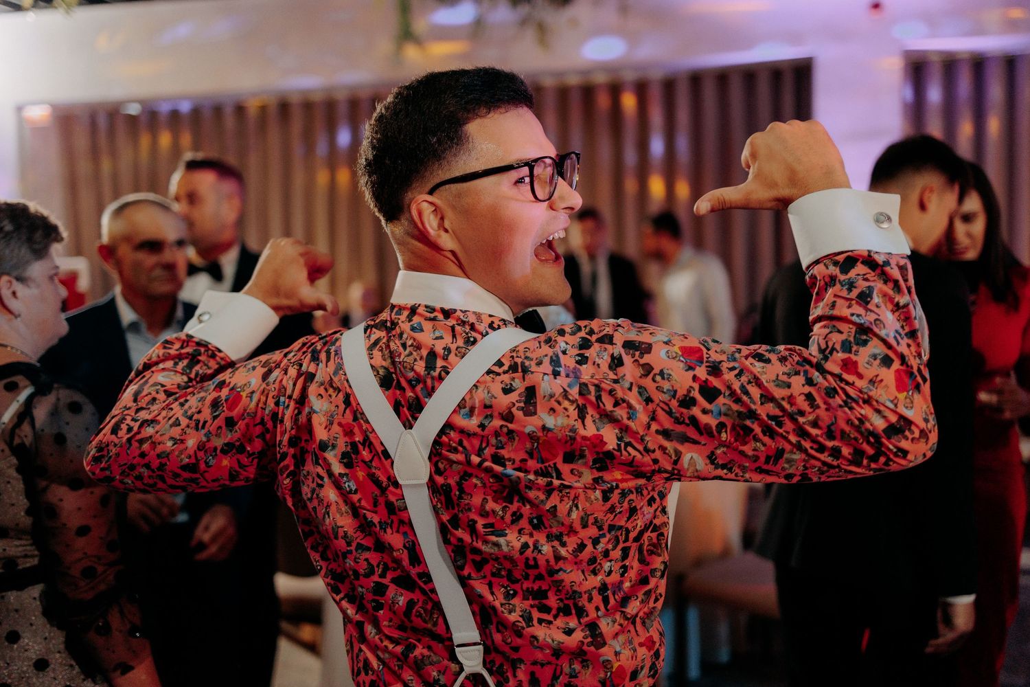 Mire purtând o cămașă cu imprimeu colorat și bretele, pozând cu spatele în timp ce dansează; atmosferă veselă și relaxată la nuntă.