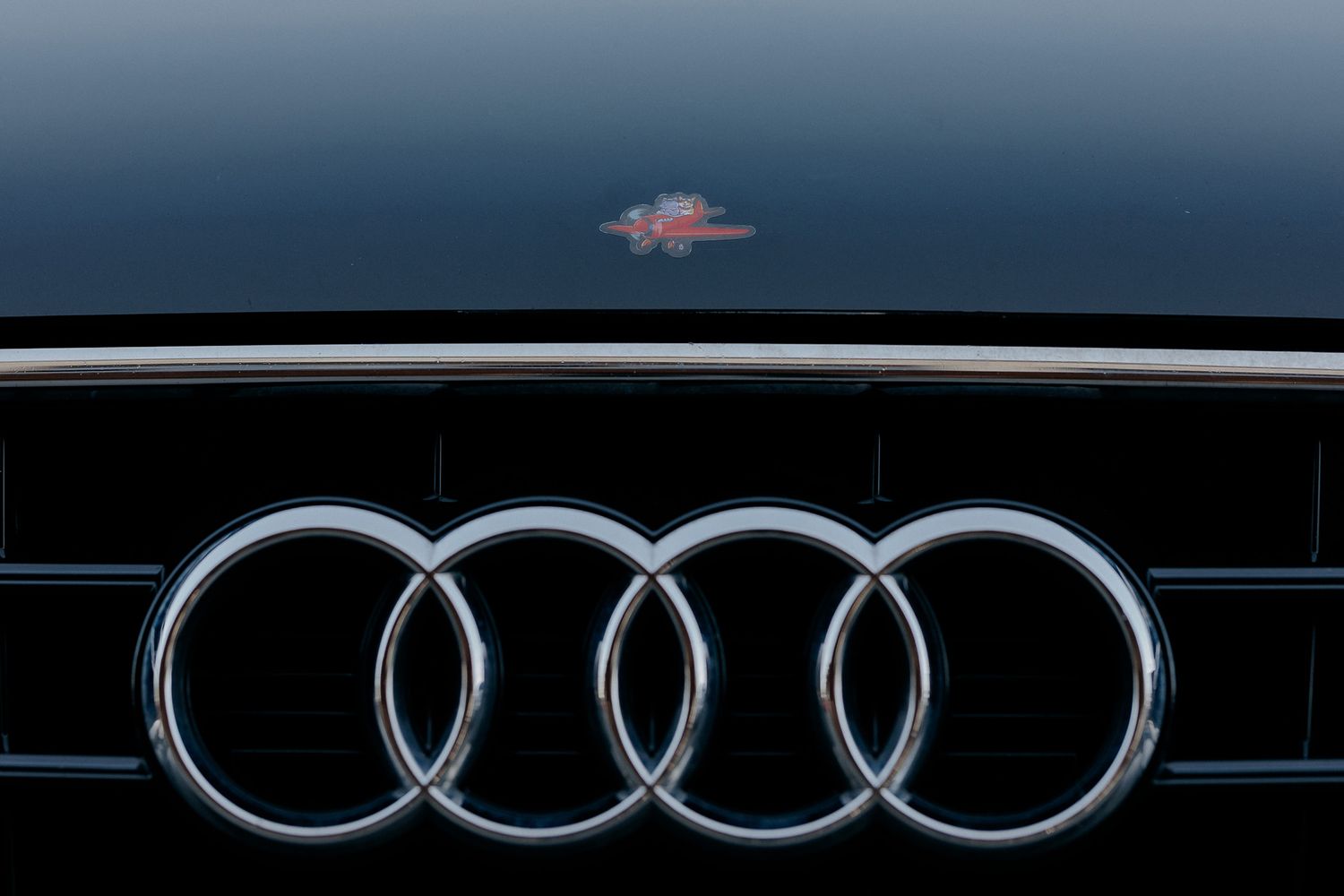 Detaliu al grilei frontale a unui automobil Audi negru, decorat cu un sticker amuzant reprezentând un avion roșu pe capotă.