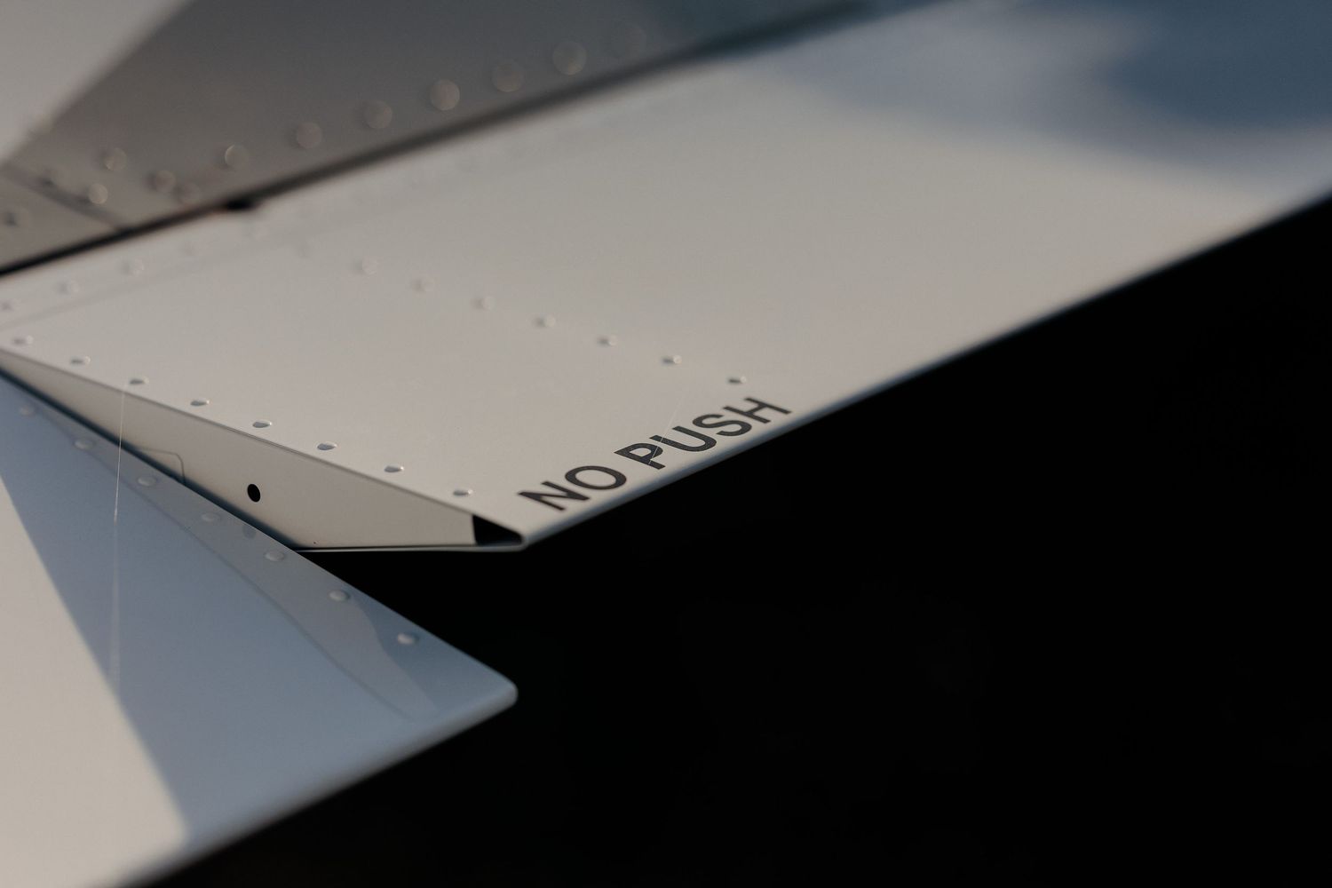 Detaliu minimalist al unei aripi de avion albă, având inscripționat mesajul de avertizare „NO PUSH” în litere negre, pe un aerodrom.