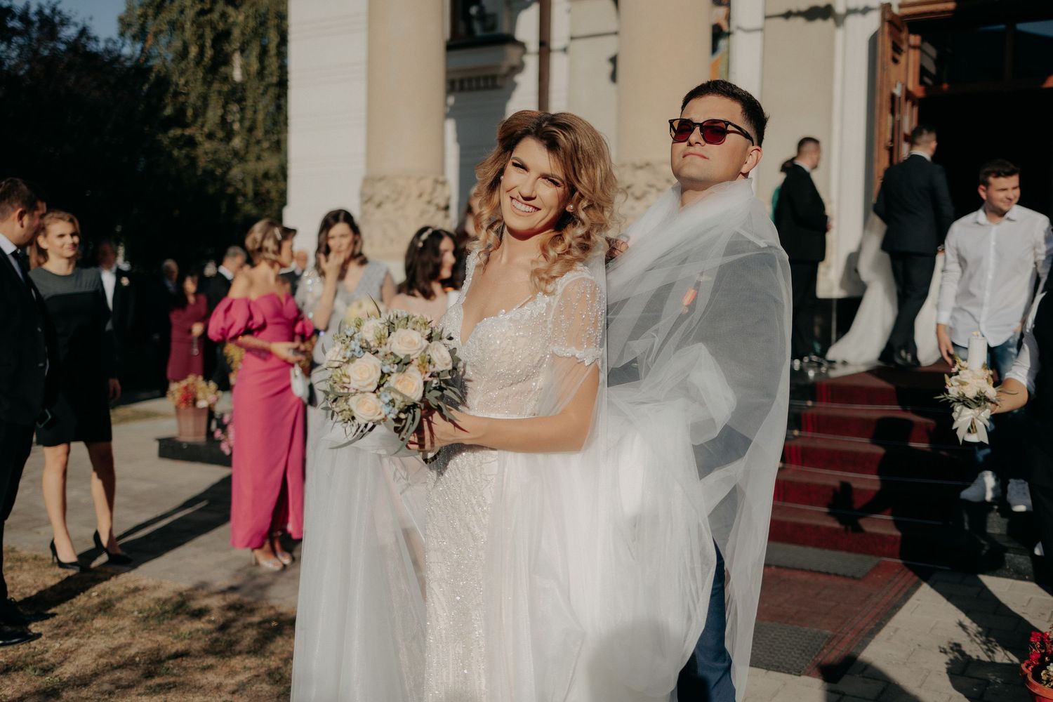 Miri zâmbind în fața bisericii după ceremonie; mirele pozează amuzant fiind înfășurat parțial în vălul lung al miresei, în lumina soarelui.