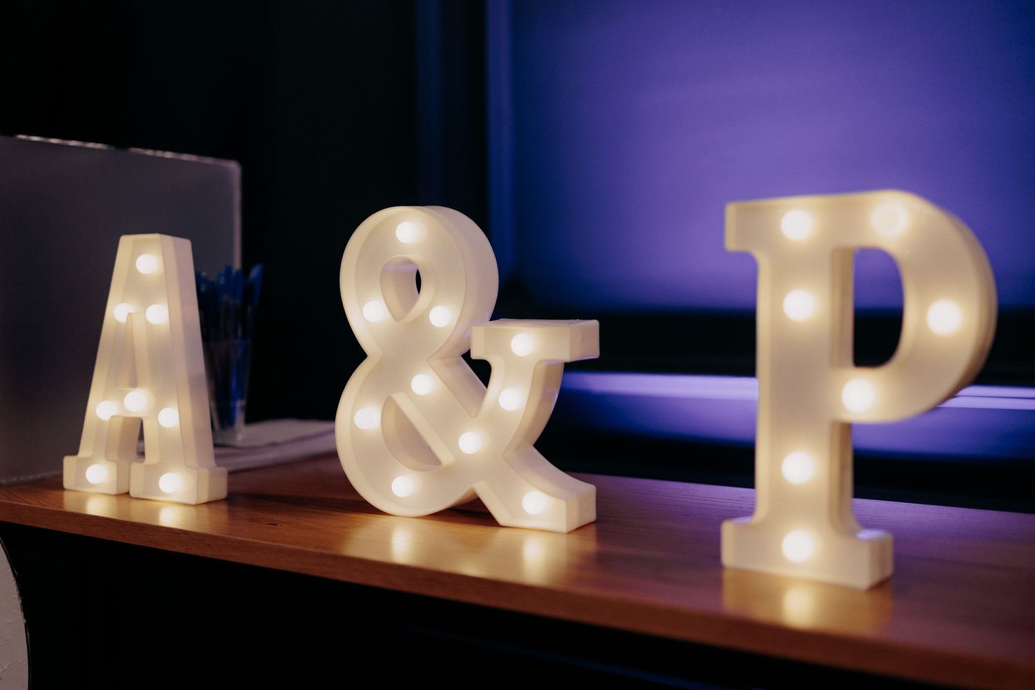 Litere volumetrice luminoase „A & P” așezate pe o poliță de lemn; detaliu decorativ romantic și personalizat pentru recepția de nuntă.
