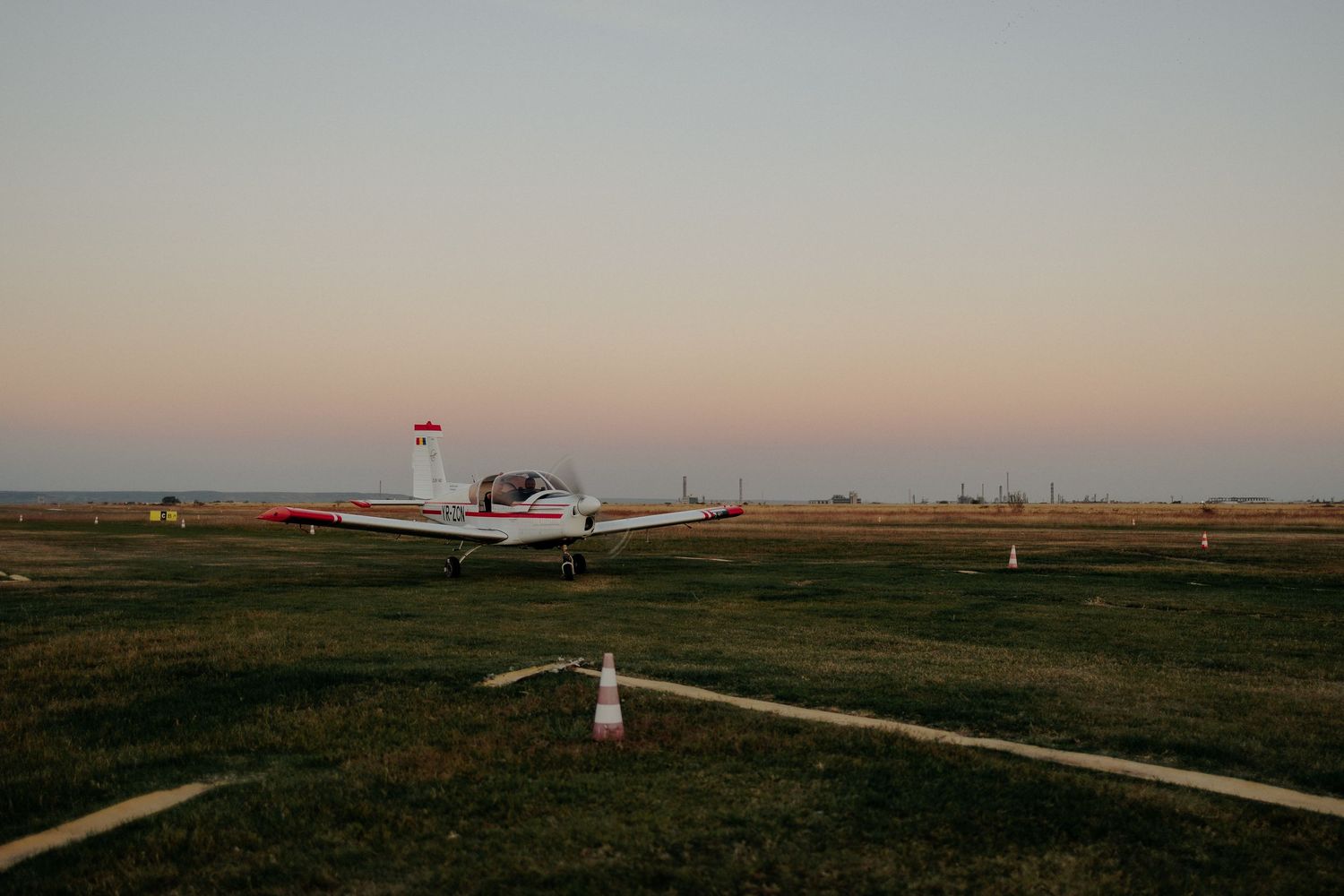 Avion utilitar alb cu detalii roșii pe un aerodrom la apus; peisaj deschis sub un cer în nuanțe calde de roz și bleu, într-o atmosferă seren
