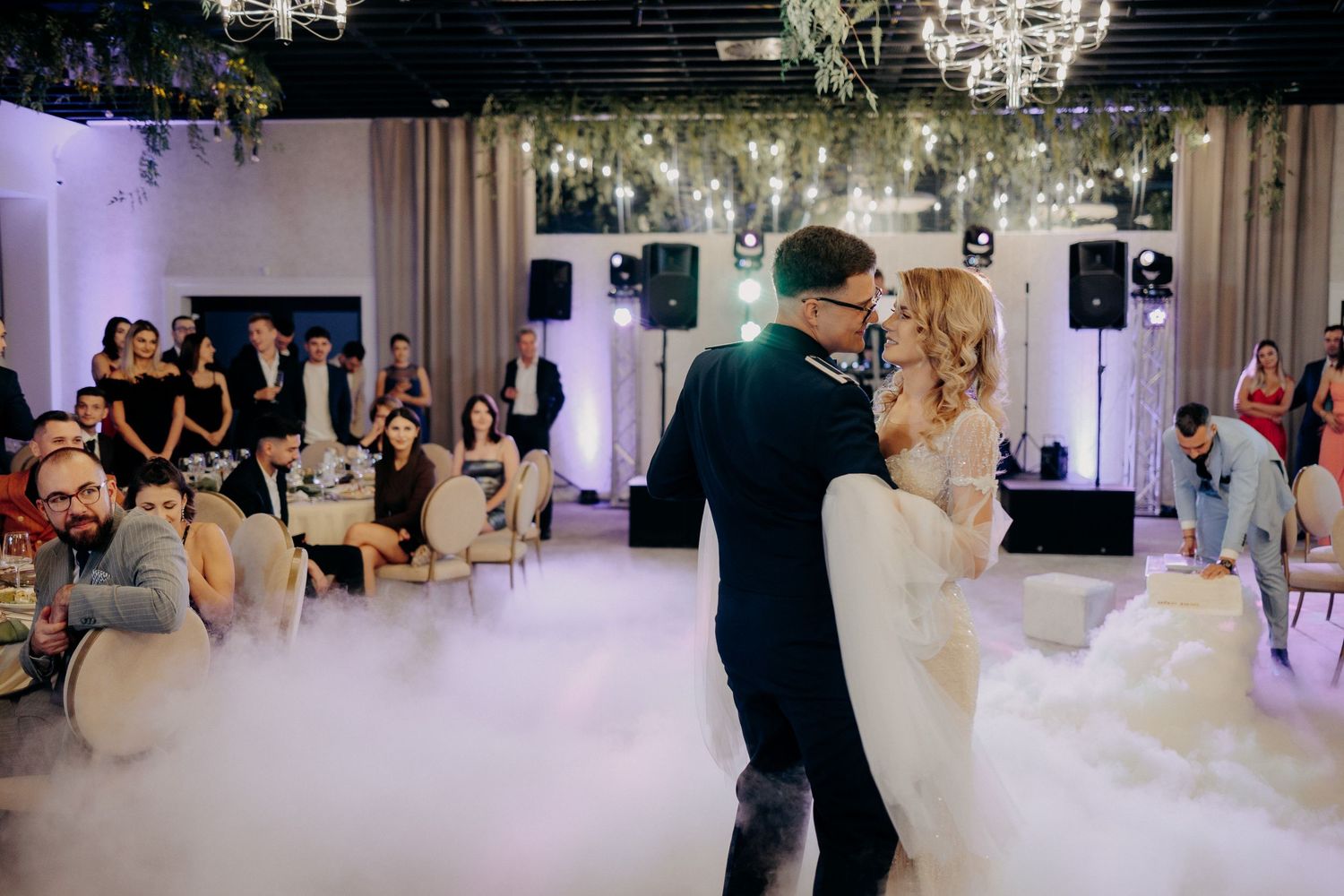 Miri dansând romantic pe ringul de dans, înconjurați de un nor spectaculos de fum alb, sub privirile invitaților la o nuntă elegantă.
