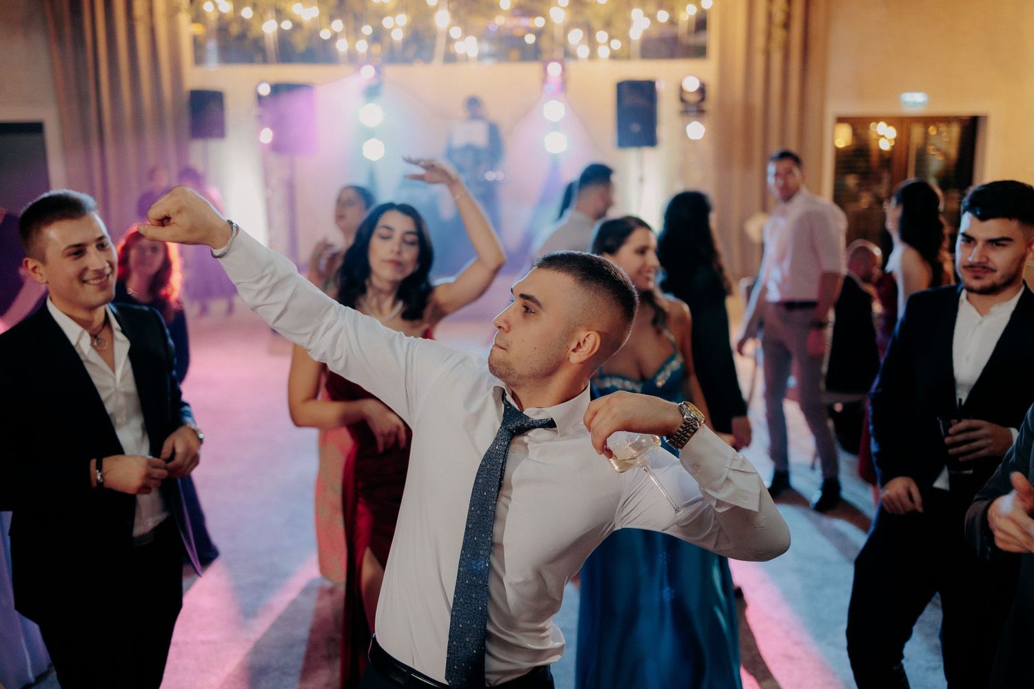Invitat dansând cu energie și ținând un pahar; moment plin de entuziasm pe ringul de dans la o petrecere de nuntă dinamică.