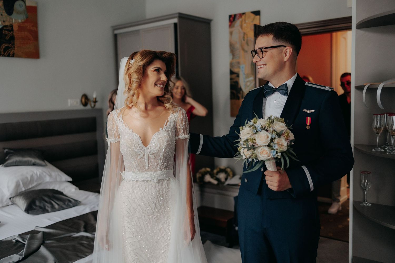 Mire și mireasă zâmbindu-și cu emoție în timpul pregătirilor; mirele în uniformă militară ține un buchet de trandafiri albi și flori bleu.