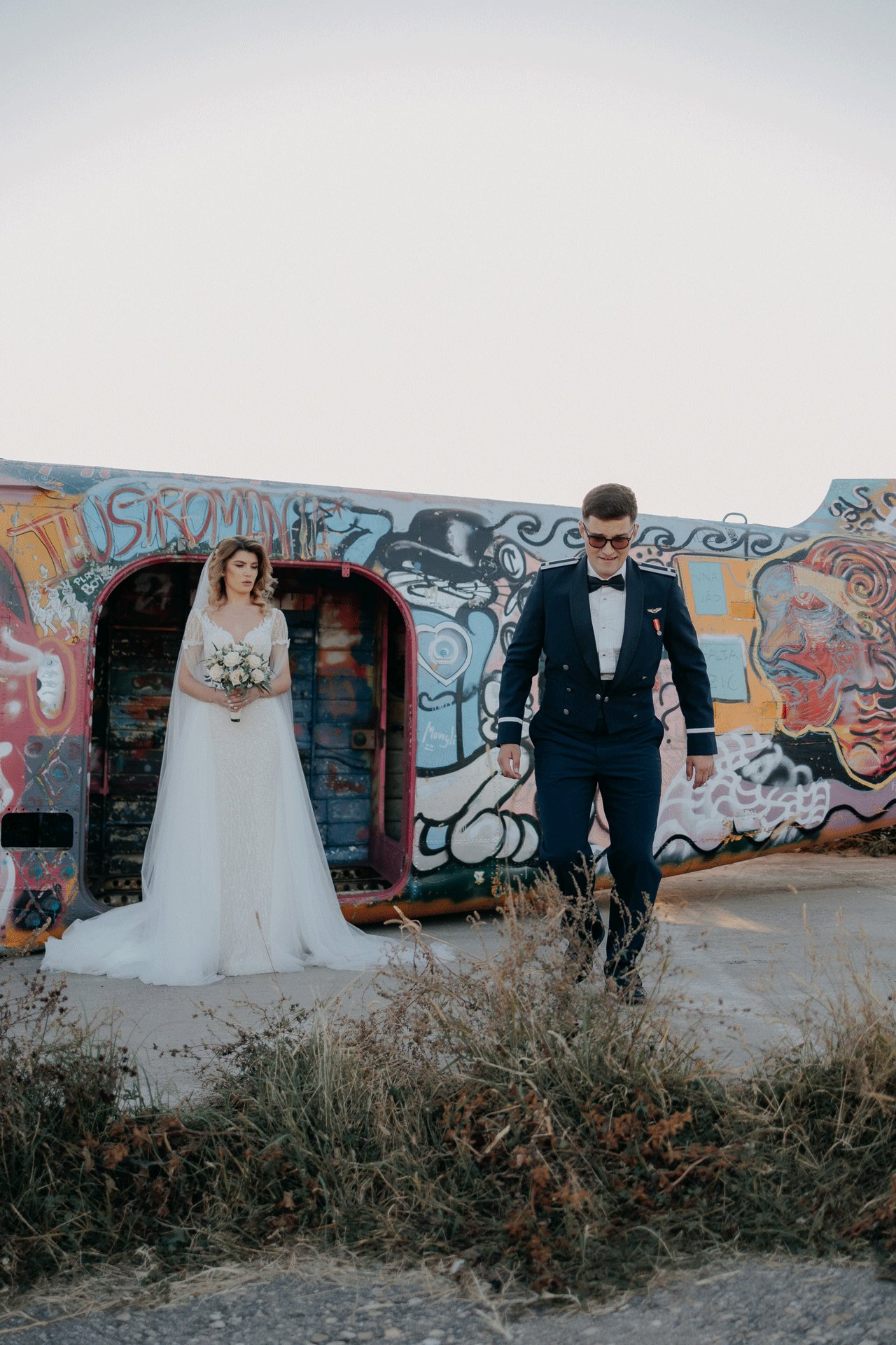 Mireasă stând elegant în ușa unui fuselaj cu graffiti, în timp ce mirele merge vesel spre cameră; moment jucăuș pe un aerodrom.