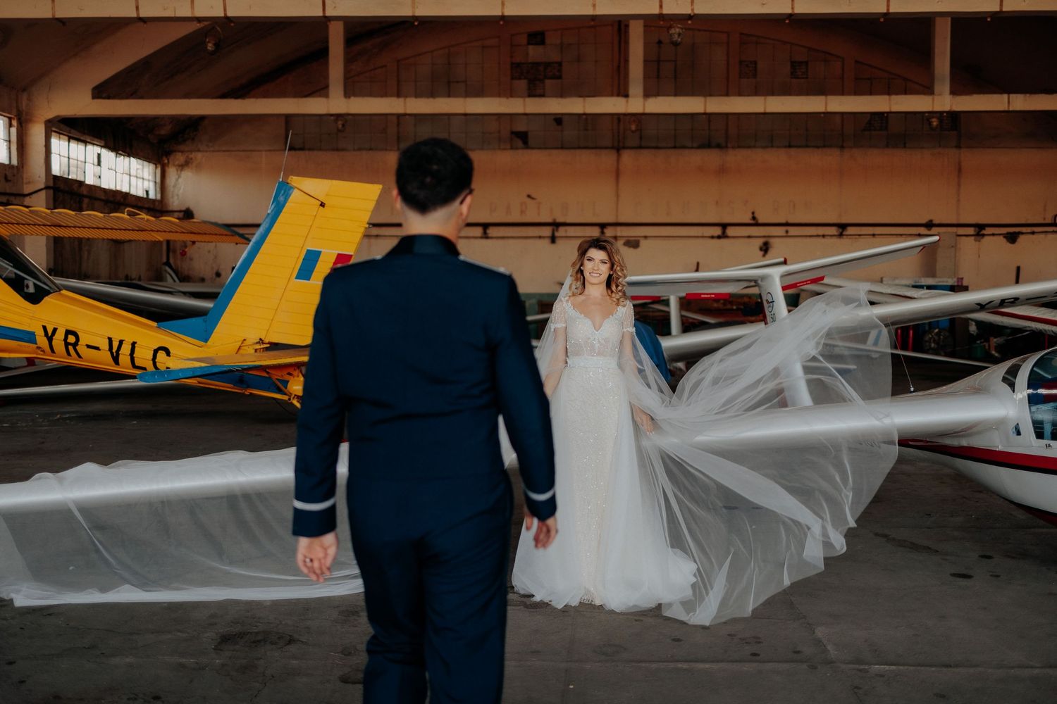 Mireasă stând în fața mirelui într-un hangar, cu vălul lung întins spectaculos pe aripile avioanelor; cadru artistic și simetric de nuntă.