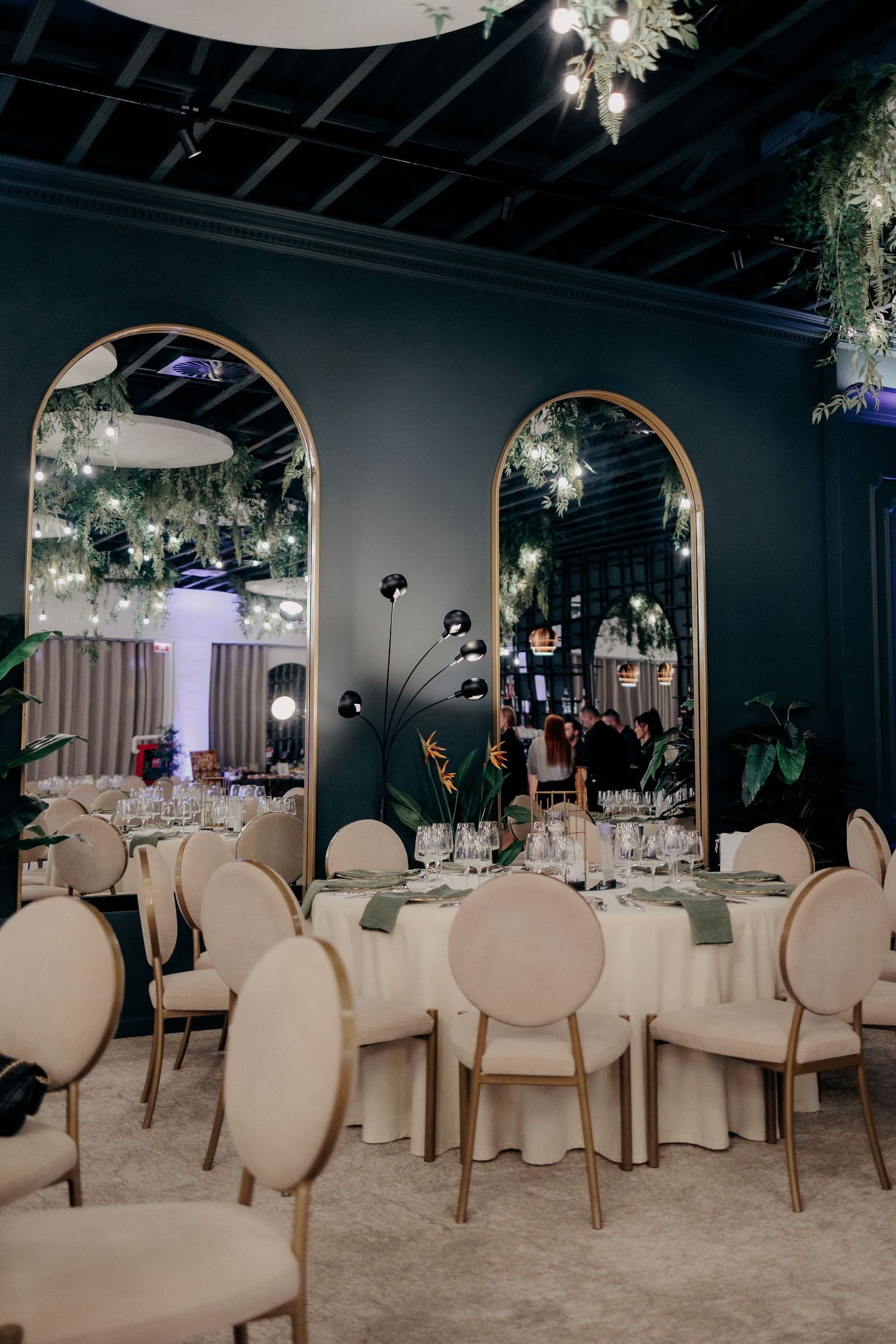 Masă rotundă de nuntă elegant decorată, reflectată în oglinzi mari boltite; design interior sofisticat cu plante verzi și lumini calde.