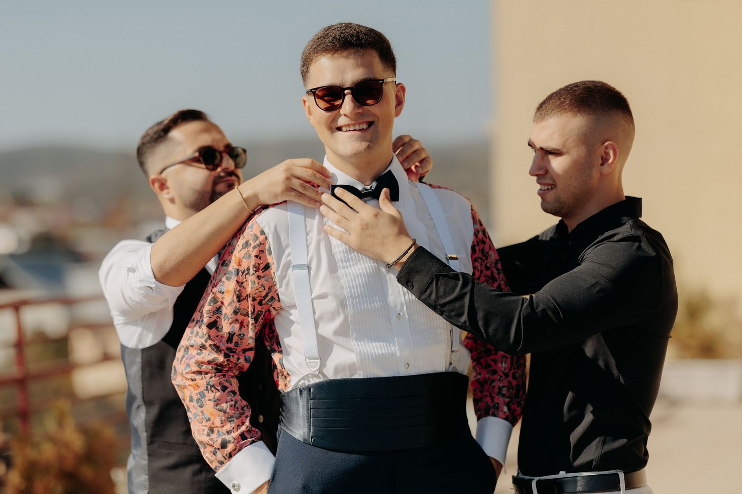 Mirele zâmbitor cu ochelari de soare ajutat de prieteni să își fixeze papionul; moment relaxat de camaraderie surprins în exterior la nuntă.