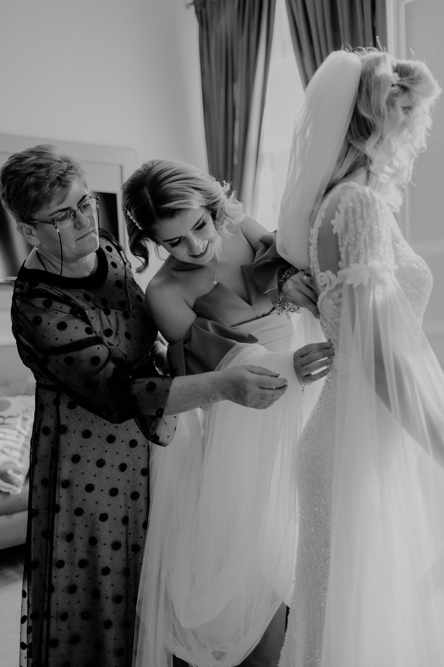 Fotografie alb-negru cu mireasa ajutată de mamă și domnișoara de onoare să îmbrace rochia; moment emoționant din timpul pregătirilor de nunt