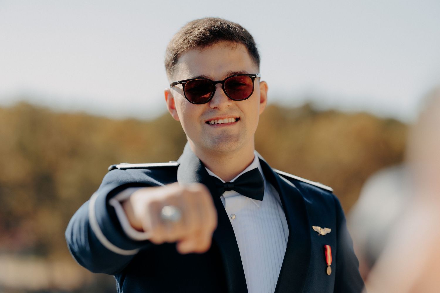 Portret cu mirele zâmbind și purtând ochelari de soare, în timp ce arată spre cameră un inel masiv; cadru relaxat în exterior la nuntă.
