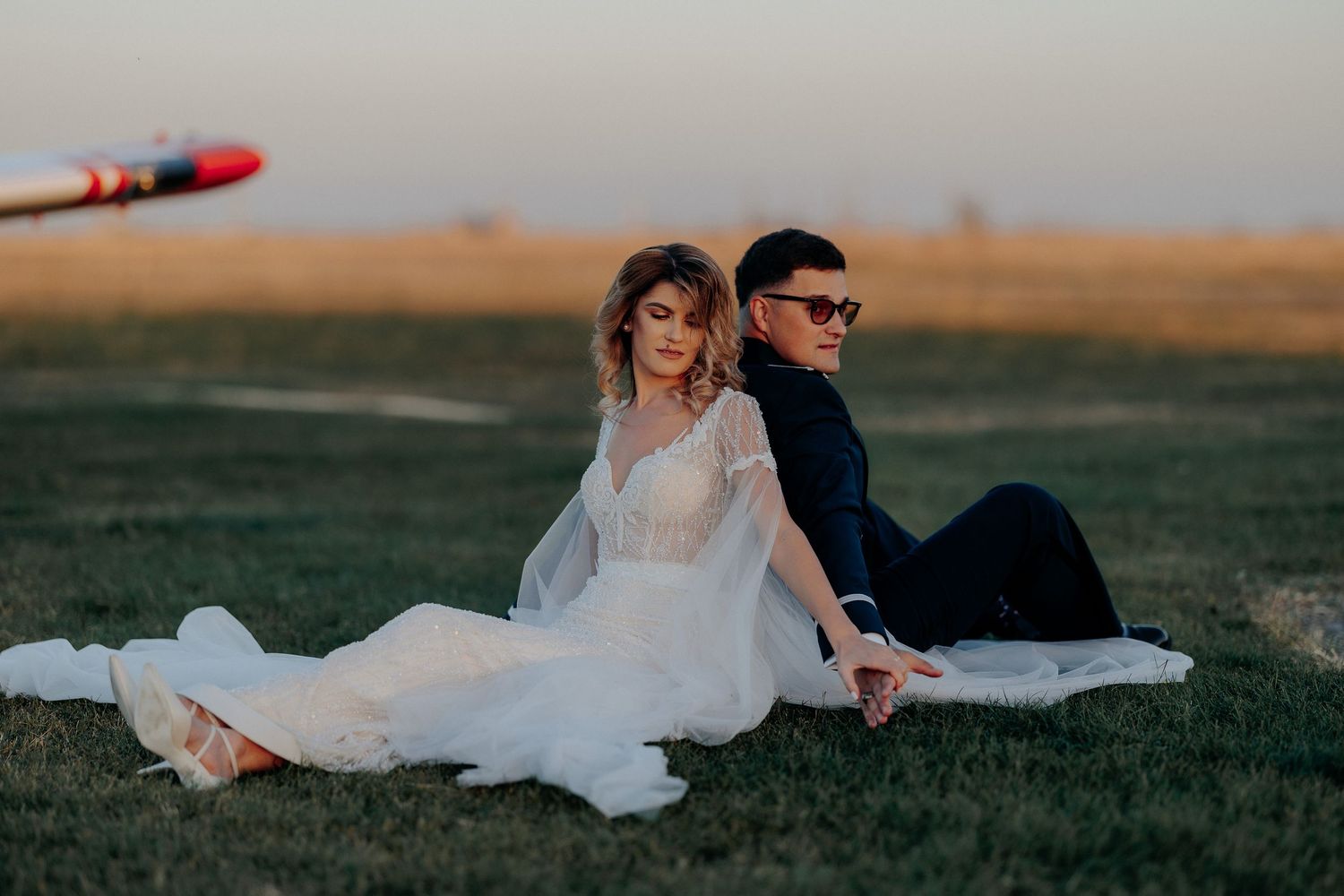 Miri stând relaxați pe iarbă, spate în spate, la apus pe un aerodrom; mirele poartă ochelari de soare, într-un cadru cinematografic.