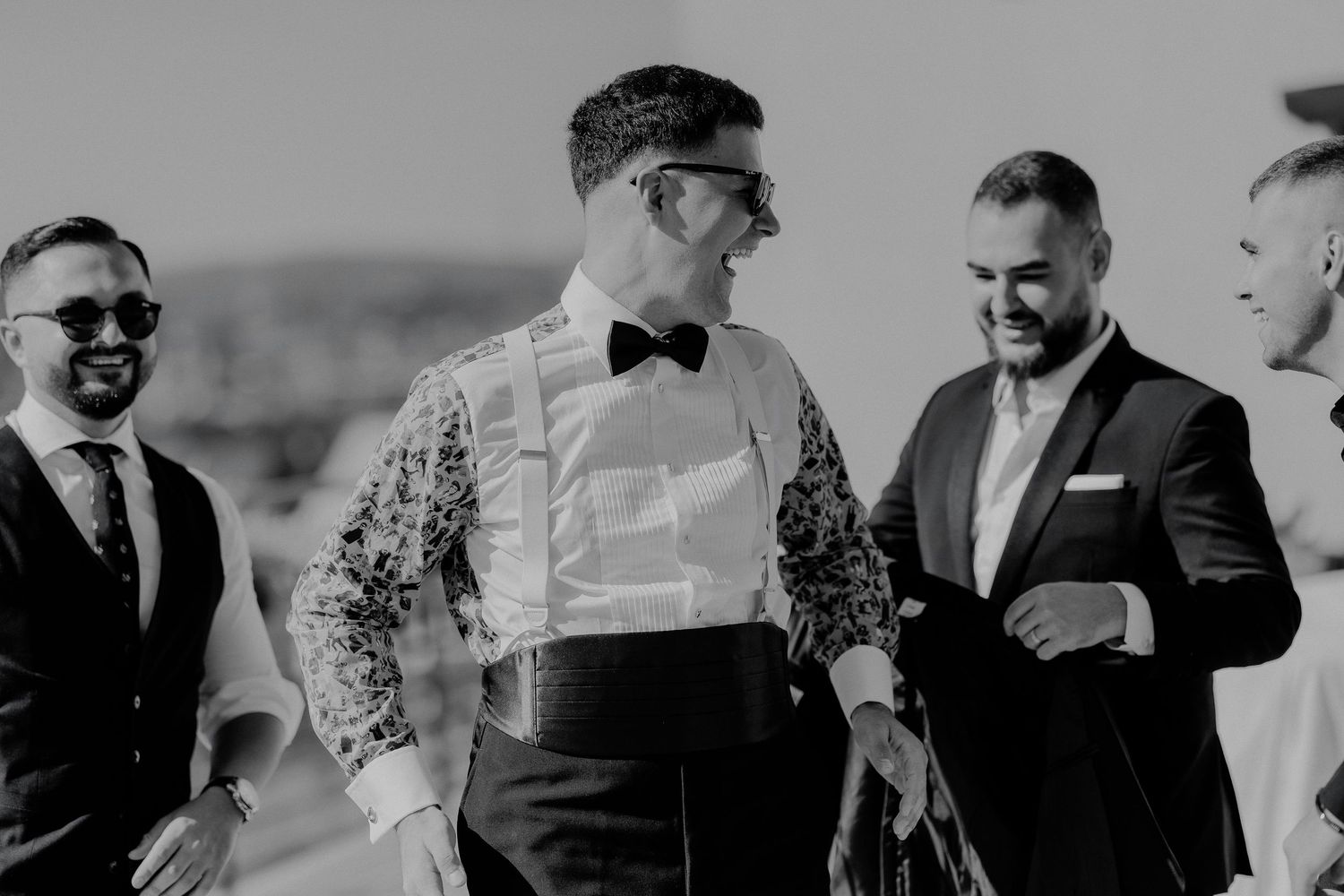 Fotografie alb-negru cu mirele râzând alături de cavalerii de onoare în exterior; moment vesel de camaraderie înaintea ceremoniei de nuntă.