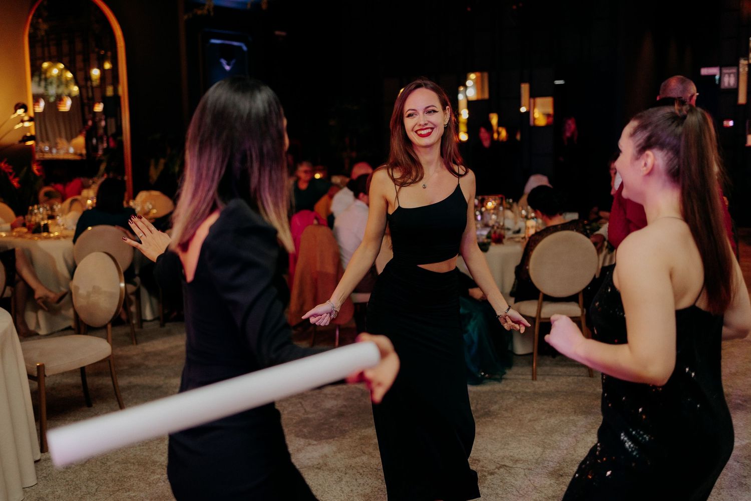 Trei invitate dansând cu entuziasm la o petrecere de nuntă; atmosferă veselă pe ringul de dans într-un decor elegant de seară.