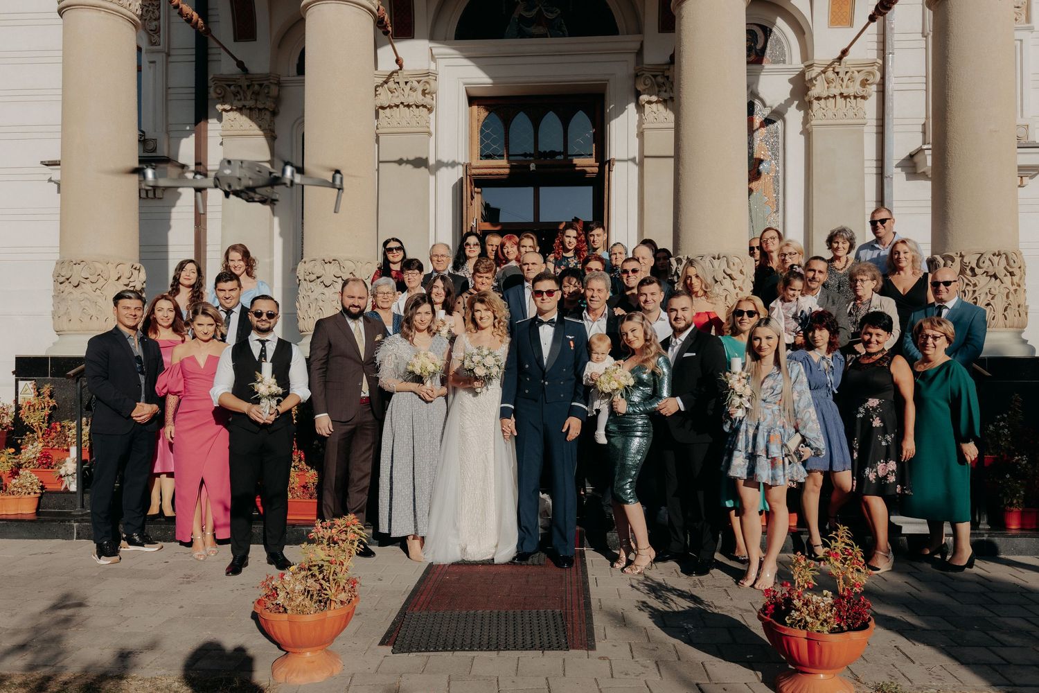 Miri și invitați pozând pe treptele unei biserici cu arhitectură clasică; un grup numeros festiv celebrând evenimentul în lumina soarelui.