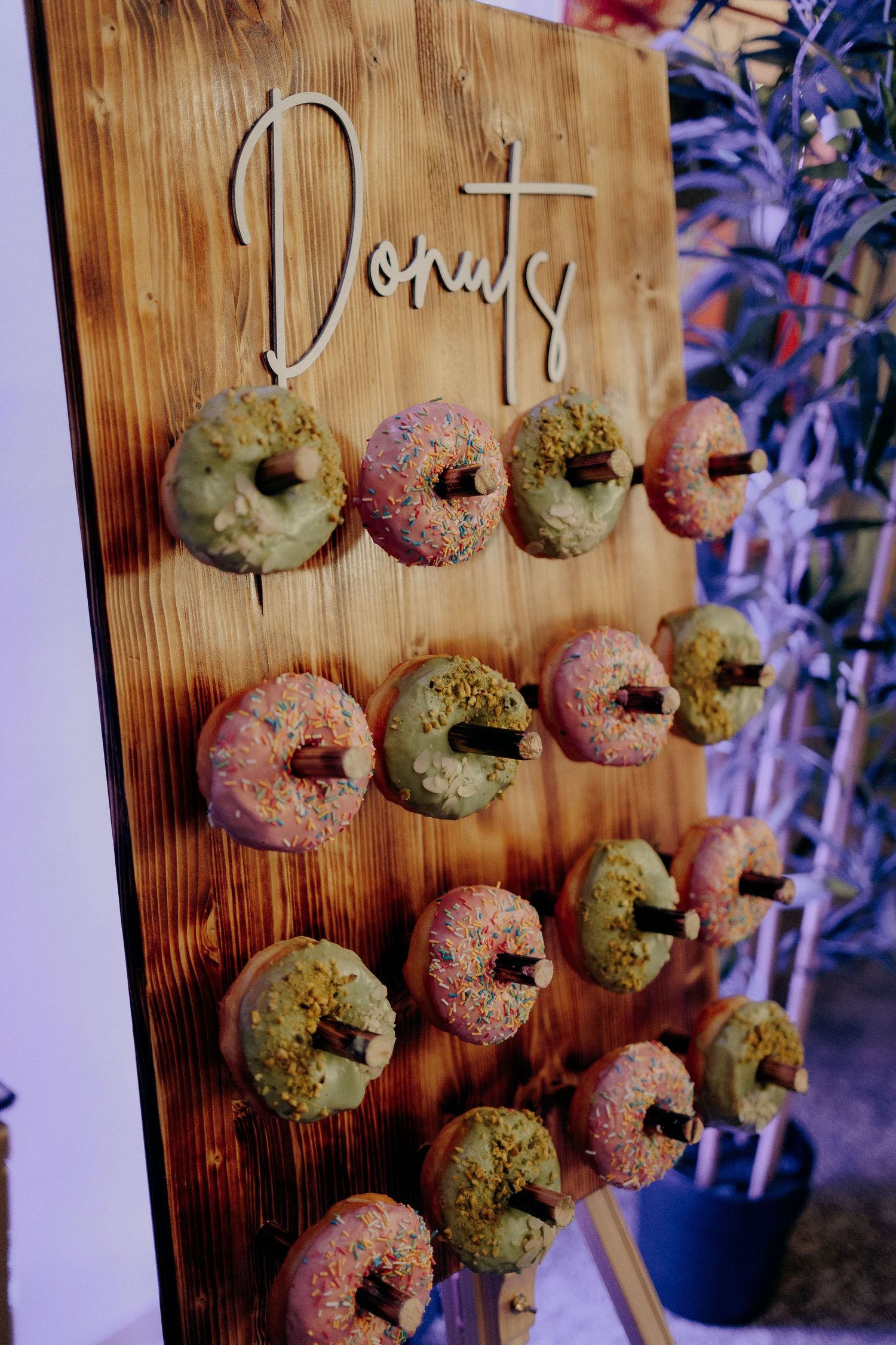 Panou din lemn cu gogoși colorate glazurate, expuse creativ pentru invitați; detaliu atractiv și delicios din zona de candy bar la nuntă.