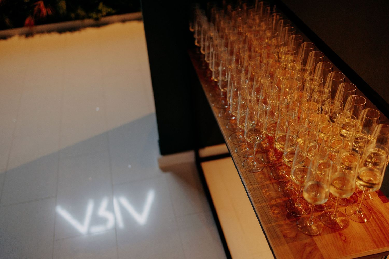 Pahare cu șampanie aliniate elegant pe un bar de lemn, luminate cald; proiecție luminoasă pe podea la intrarea în sala de nuntă.