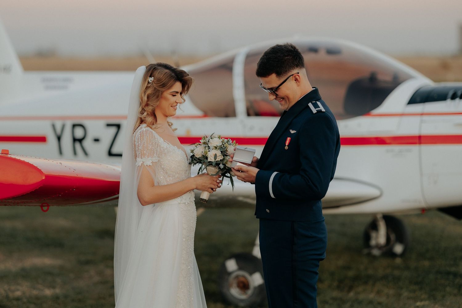 Mireasa oferindu-i mirelui o cutie de bijuterii lângă un avion; moment emoționant și zâmbete în timpul sesiunii foto pe aerodrom.