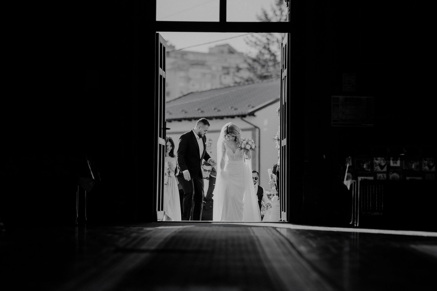 Silueta miresei și a nașului intrând în biserică; fotografie alb-negru artistică realizată din interior, cu lumina soarelui în fundal.