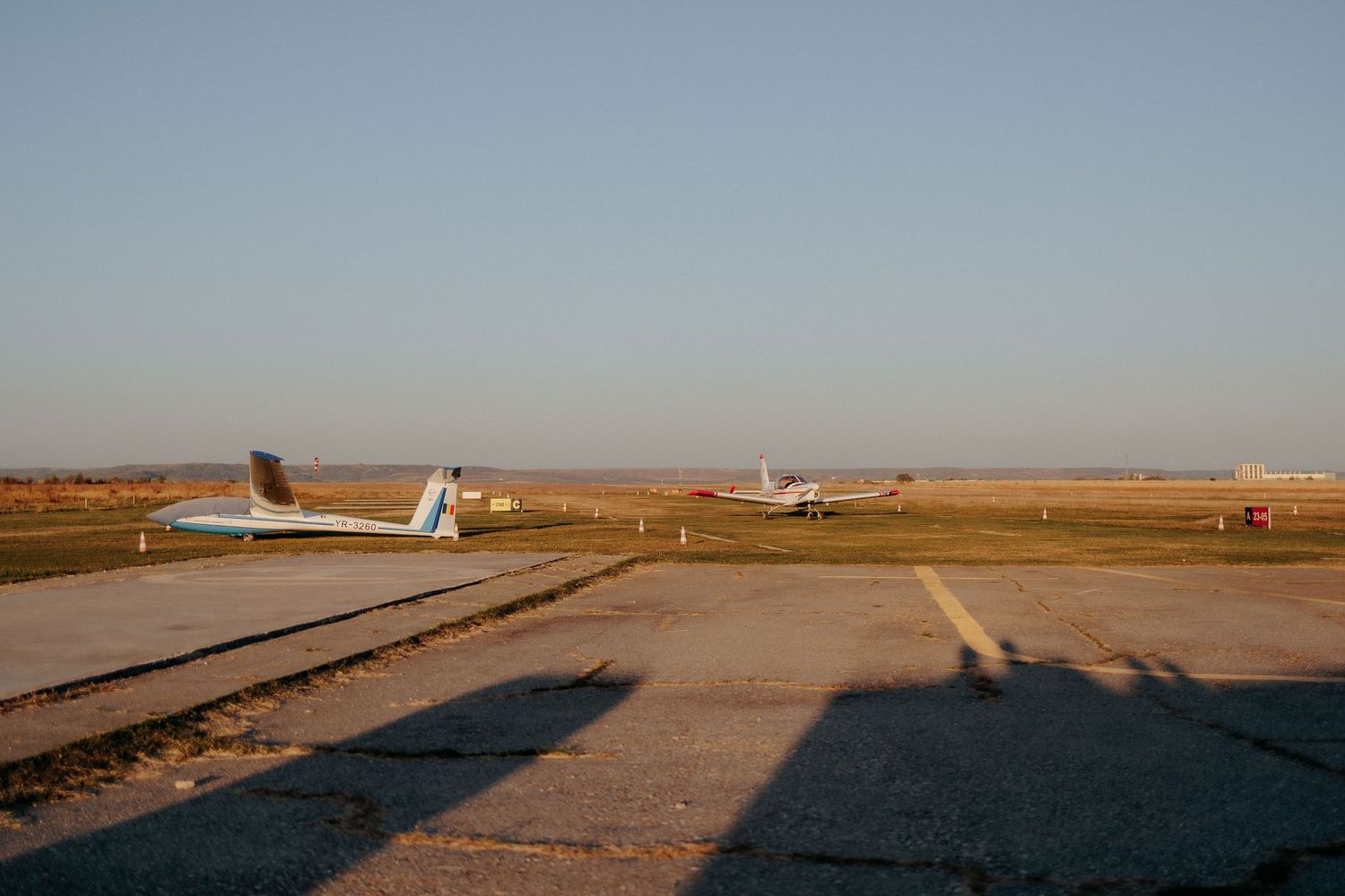 Pistă de decolare cu un avion utilitar și un planor sub lumina caldă a soarelui; peisaj vast de aerodrom într-o după-amiază de toamnă.
