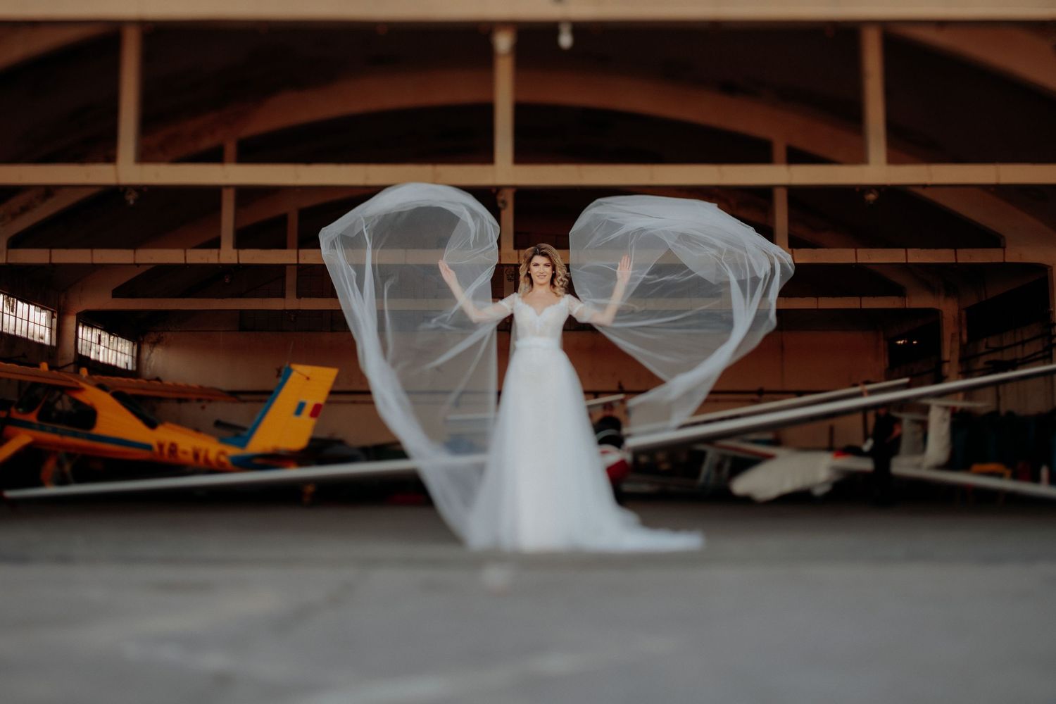 Mireasă în centrul unui hangar, ridicându-și brațele pentru a face vălul lung să plutească; cadru artistic și simetric cu avioane în fundal.