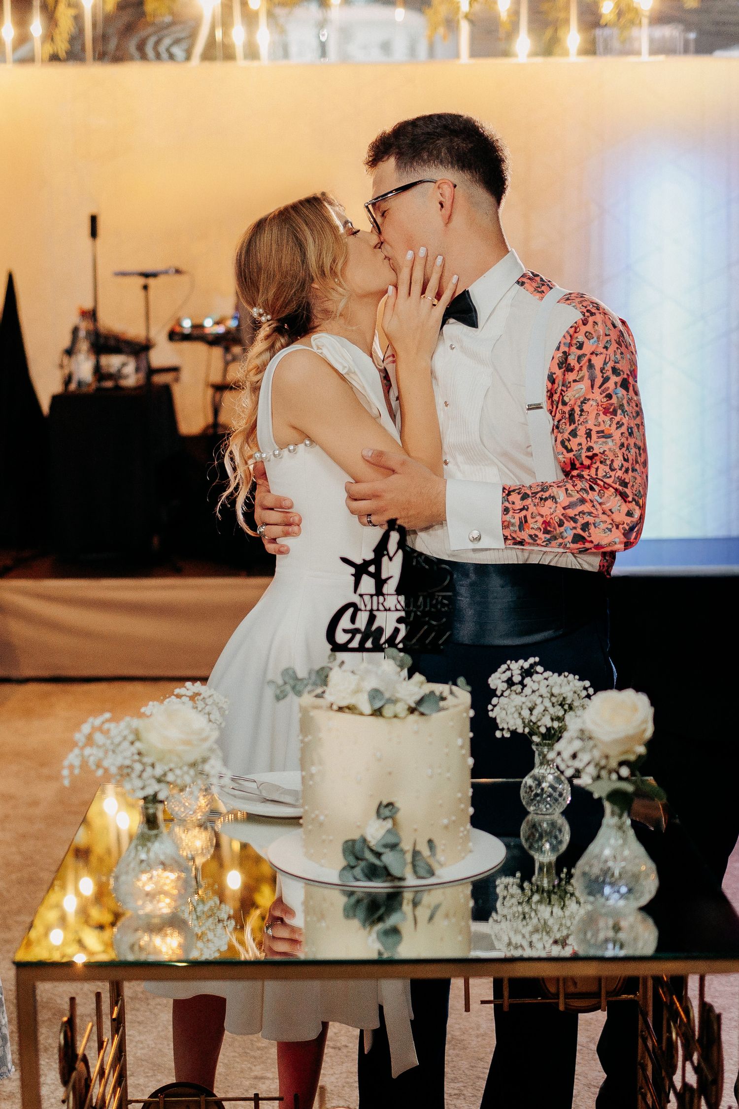 Alina și Petruț se sărută în spatele tortului de nuntă; moment festiv cu decor elegant, lumini calde și topper personalizat pe tort.
