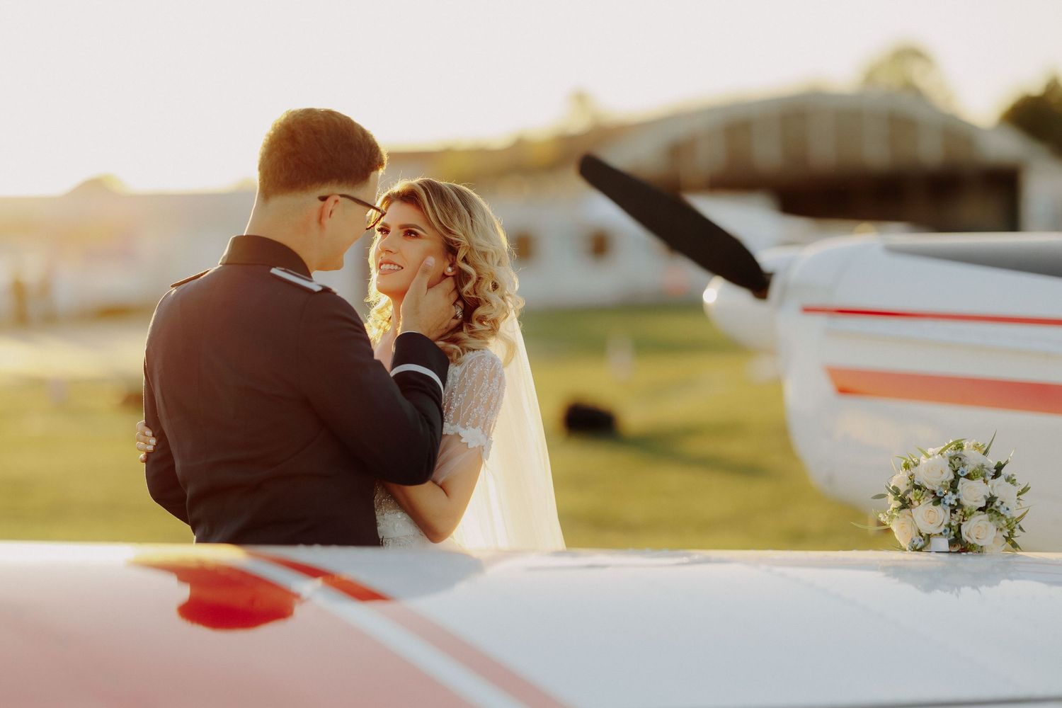 Mire privind-o cu dragoste pe mireasă și mângâindu-i fața pe un aerodrom; portret romantic la apus, cu un avion și un buchet în fundal.