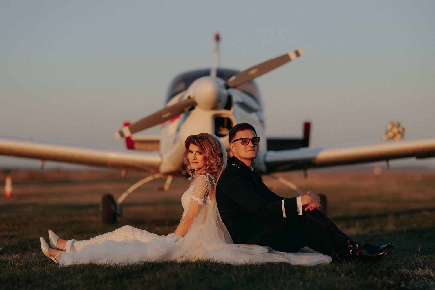 Miri stând spate în spate pe iarbă, direct în fața unui avion, sub lumina caldă a apusului; cadru simetric și elegant pe un aerodrom.