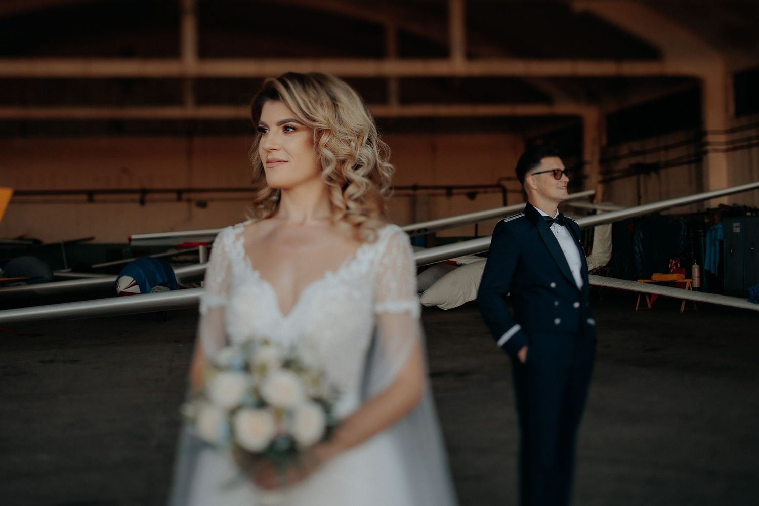 Portret al miresei zâmbitoare cu un buchet în prim-plan; mirele este vizibil în fundal, amândoi aflându-se într-un hangar de avioane.