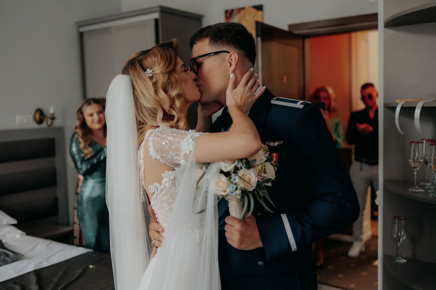Mire și mireasă sărutându-se cu emoție în timpul pregătirilor; mirele în uniformă militară ține un buchet de trandafiri albi și flori bleu.