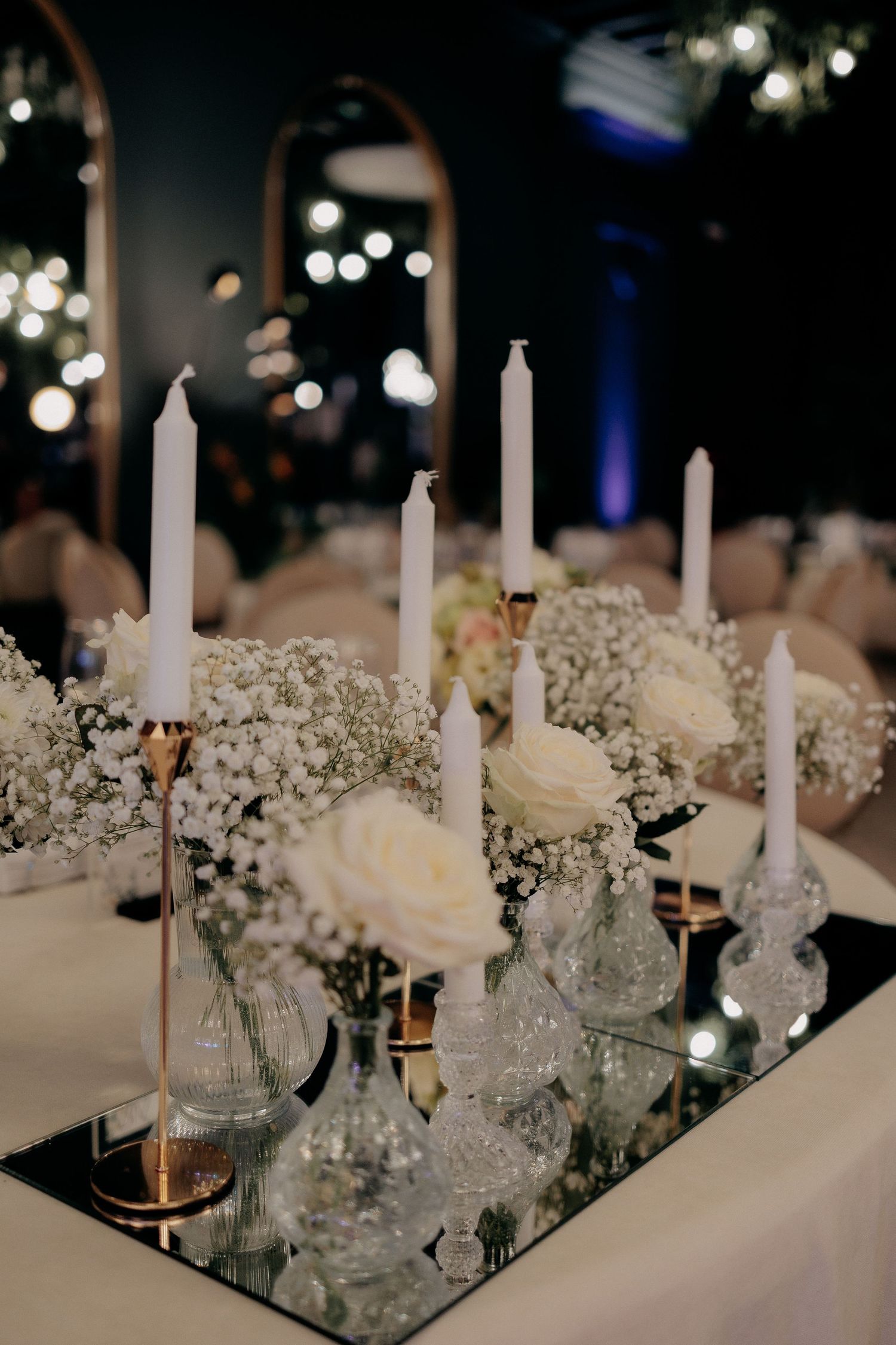 Aranjament elegant de masă cu trandafiri albi, gypsophila și lumânări subțiri în suporturi de cristal, așezate pe o bază de oglindă la nuntă