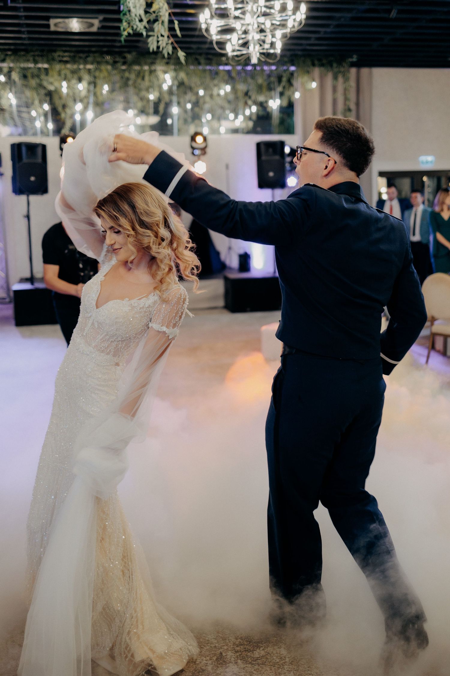 Mireasă elegantă dansând pe ringul cuprins de fum alb, în timp ce mirele o rotește delicat; moment magic și rafinat la petrecerea de nuntă.