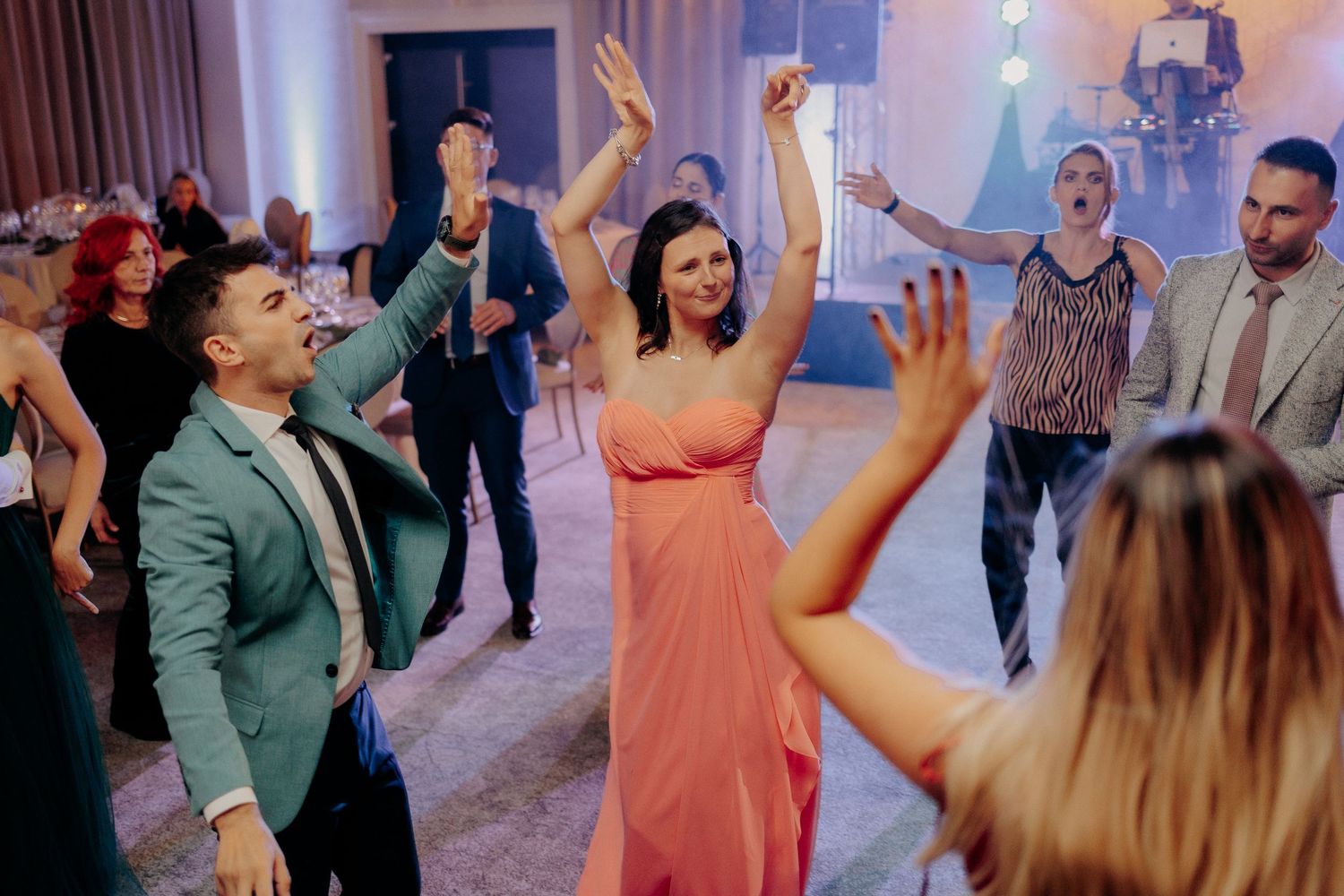 Invitată în rochie lungă corai dansând cu brațele ridicate; atmosferă plină de viață și invitați veseli pe ringul de dans la nuntă.