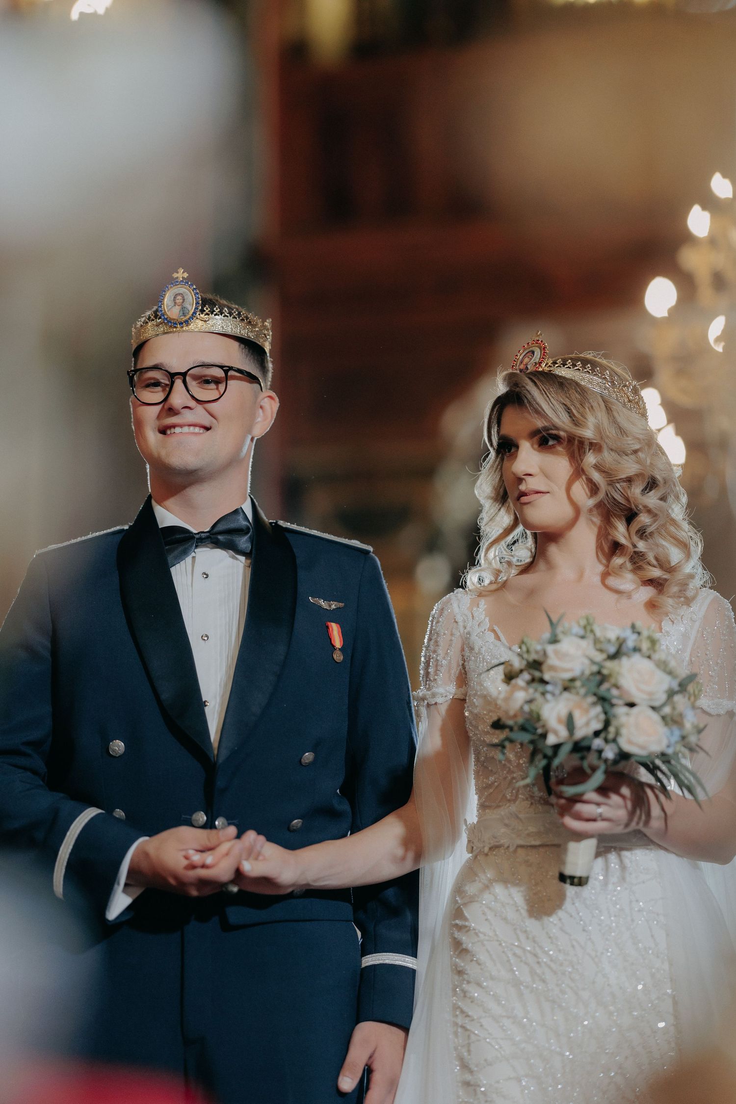 Portret cu mirii purtând coroane în timpul cununiei religioase; mirele în uniformă zâmbește, iar mireasa ține un buchet de trandafiri albi.
