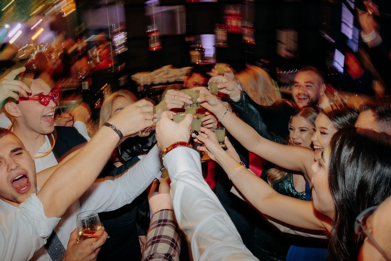 Grup vesel de tineri ciocnind shot-uri la barul unei nunți; atmosferă de petrecere efervescentă și momente pline de entuziasm.