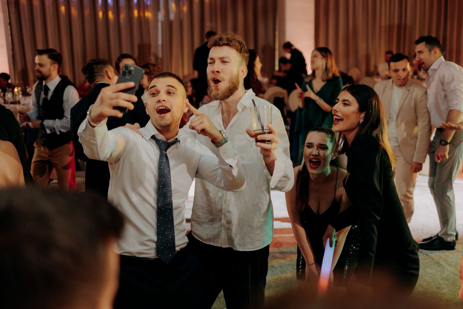 Grup de tineri făcându-și un selfie și cântând cu entuziasm pe ringul de dans, surprinzând bucuria petrecerii de nuntă.