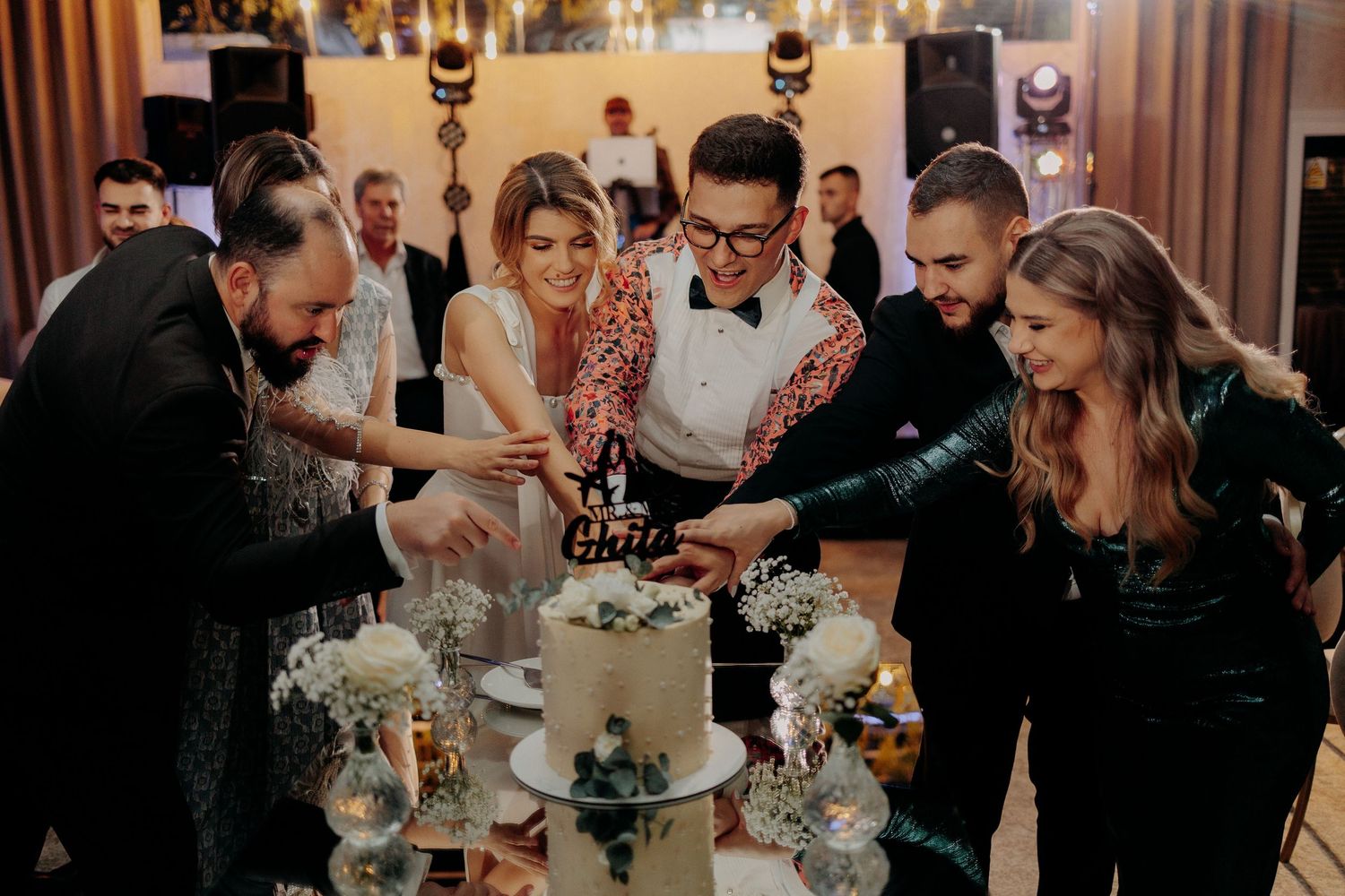 Alina, Petruț și prietenii tăind tortul de nuntă; moment plin de veselie și colaborare la petrecere, într-un decor elegant cu lumini calde.