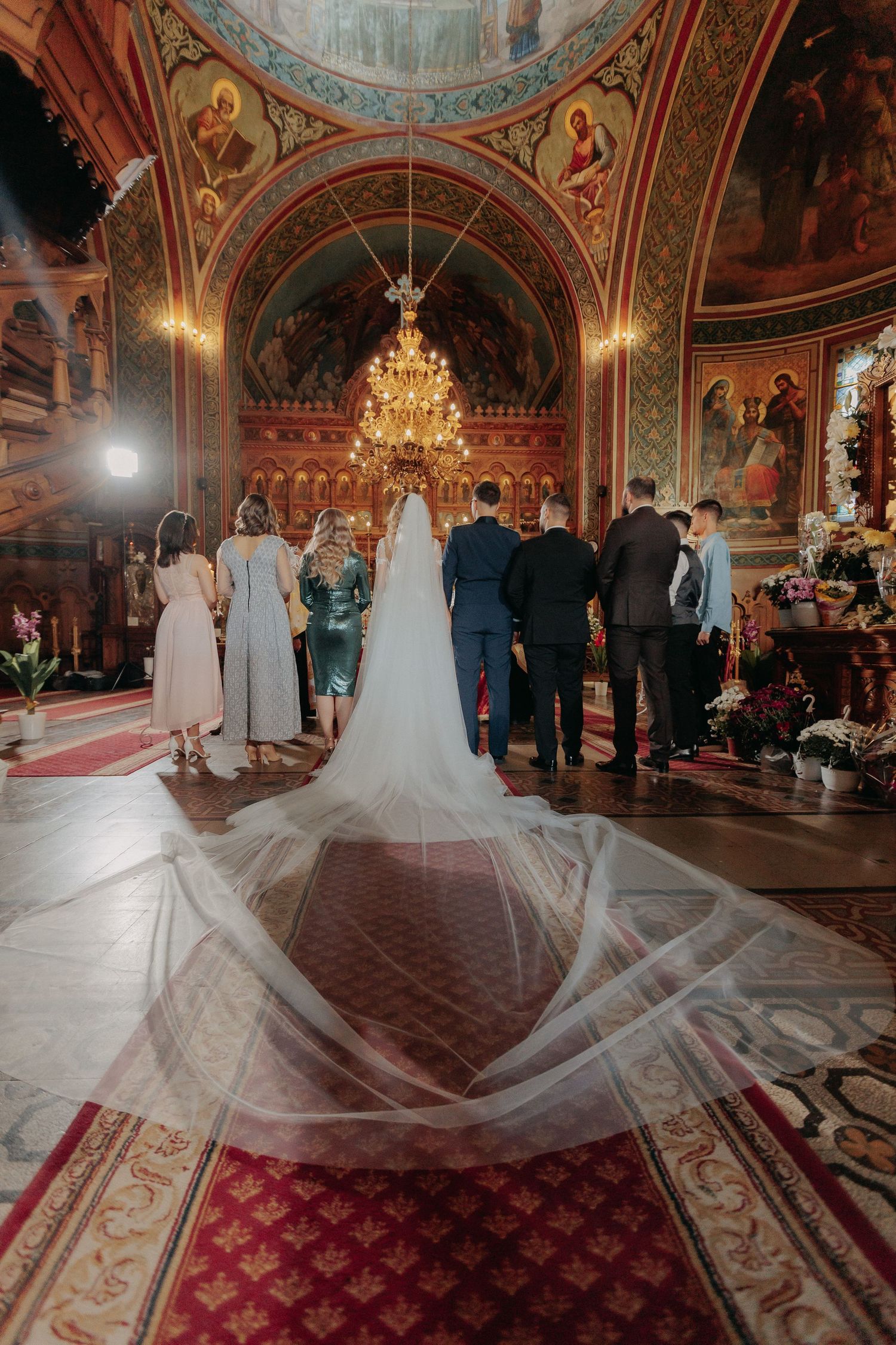 Vedere din spate cu trena lungă a miresei întinsă spectaculos pe covorul bisericii în timpul ceremoniei; mirii stau în fața altarului.