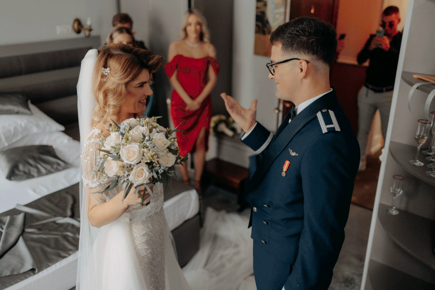 Mireasă și mire în uniformă militară privindu-se cu dragoste în timpul pregătirilor; moment emoționant cu invitați în fundal.