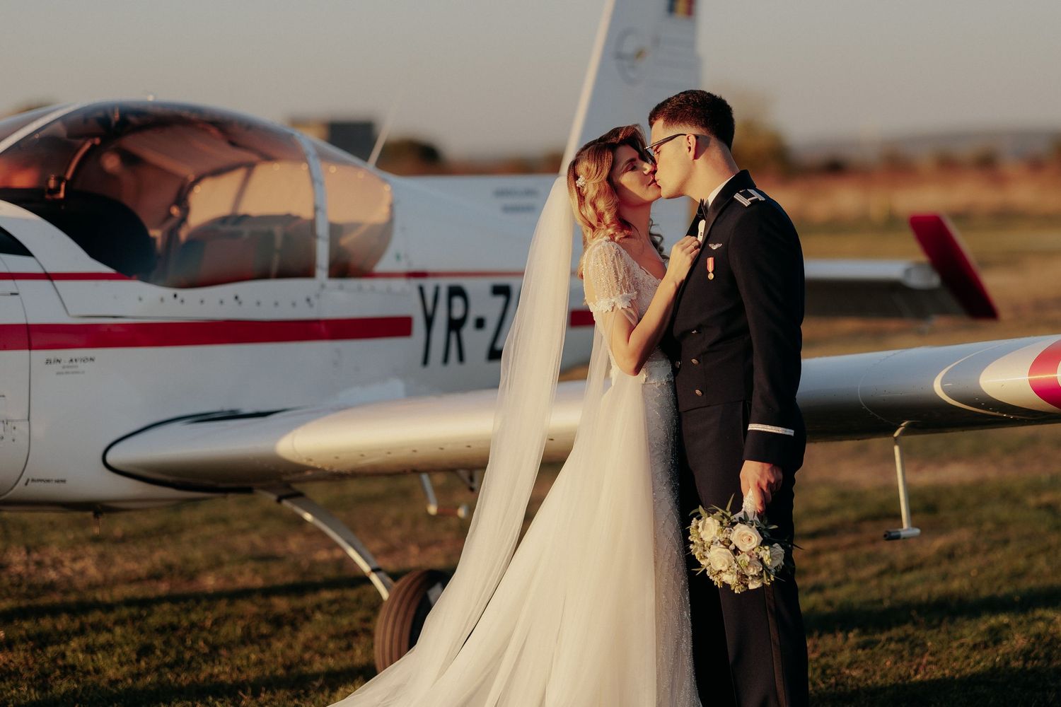 Miri sărutându-se romantic lângă un avion la apus; mireasa are un văl lung și un buchet alb, într-un cadru elegant pe un aerodrom.