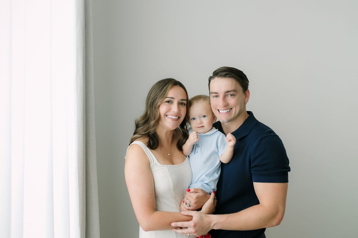 A smiling young family with a mother, father, and baby boy posing together in a bright, minimalist studio setting.